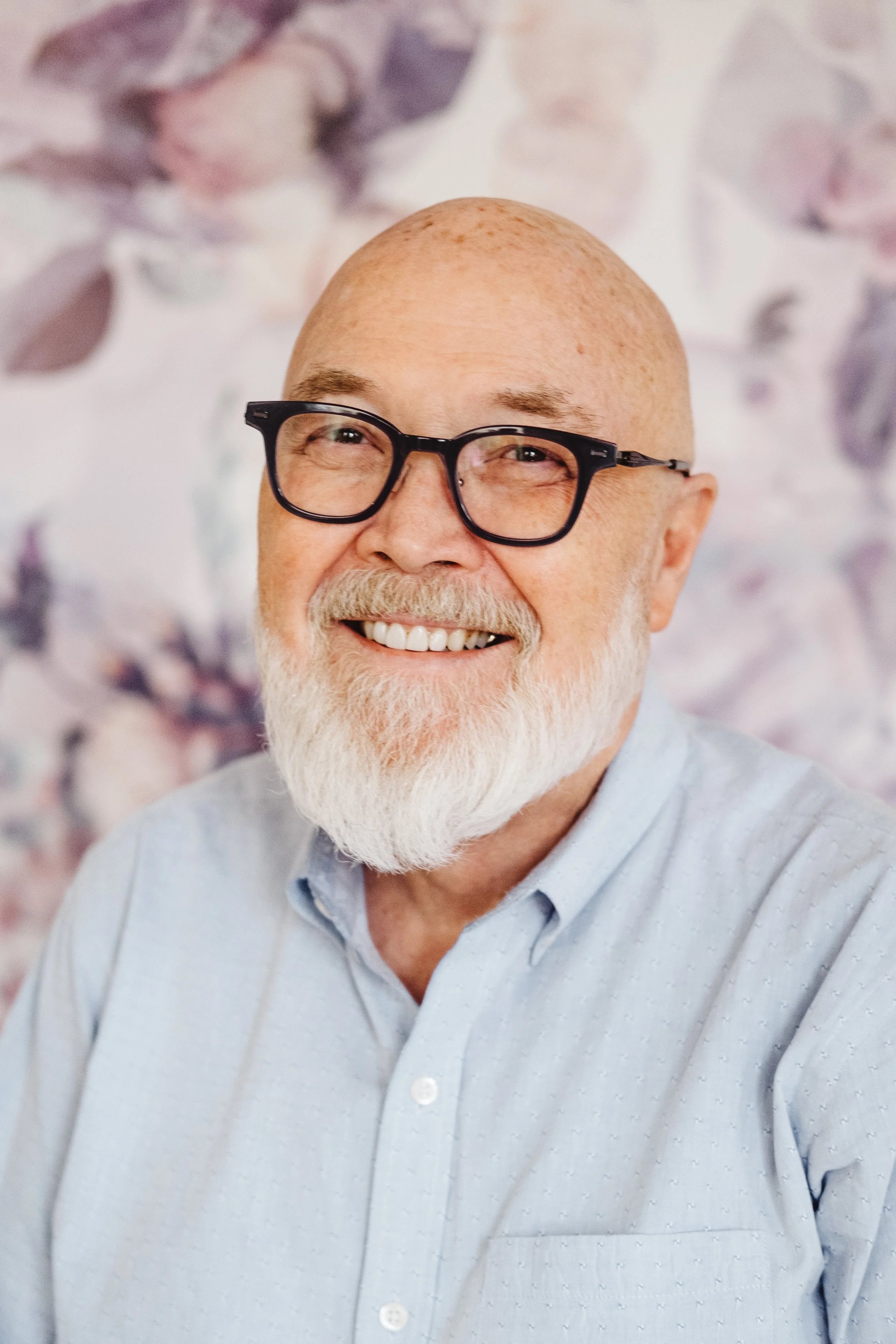 A smiling elderly man with a white beard, wearing glasses and a light blue button-up shirt, standing in front of a blurred floral background.