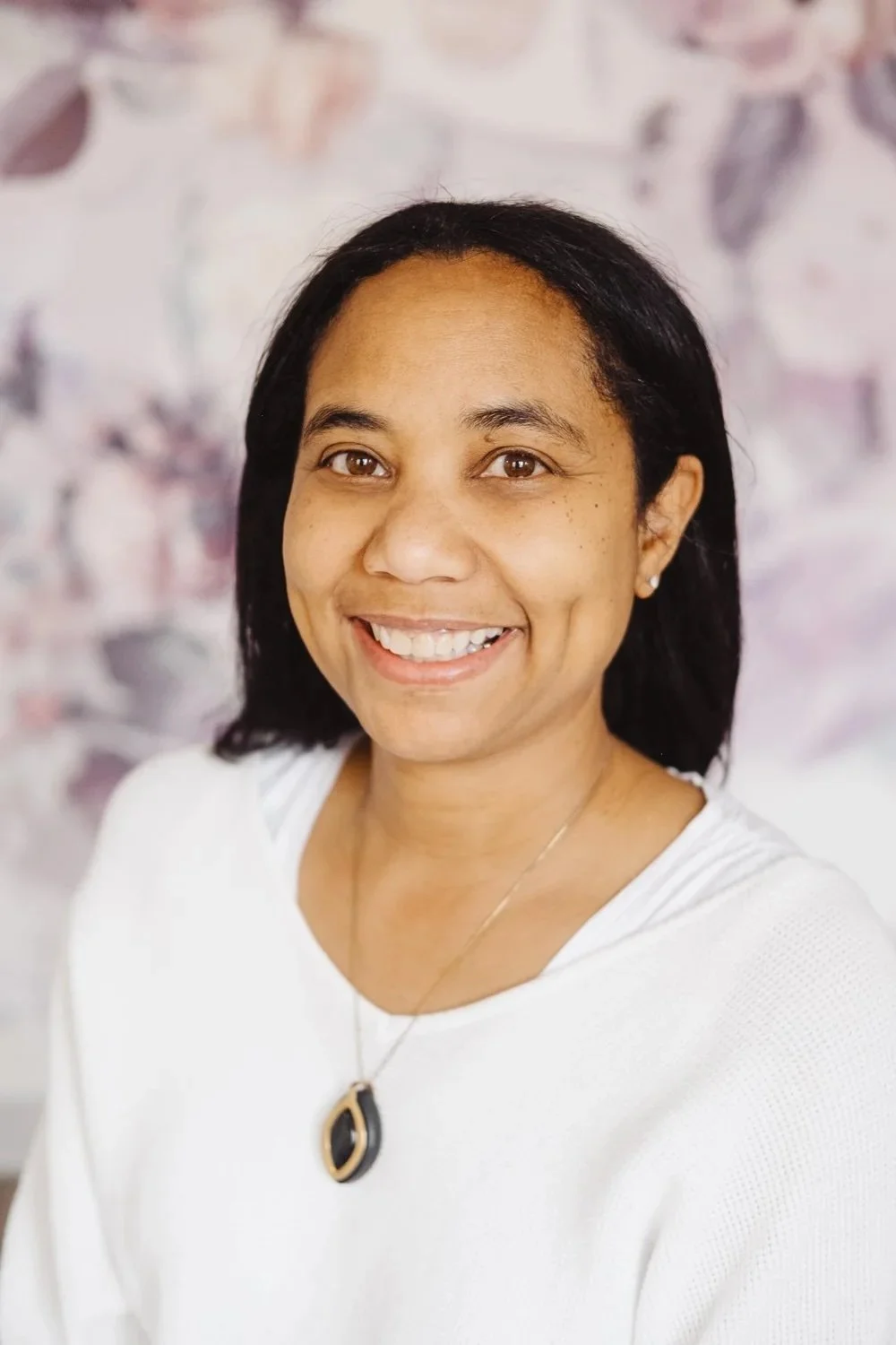 A smiling woman with dark hair, wearing a white top and a black and gold necklace, posing in front of a blurred floral background.
