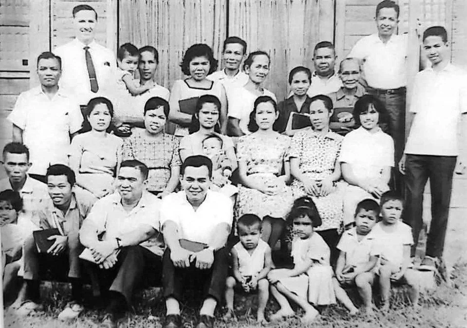 A black and white photograph of a large group of people, including men, women, and children, posing outdoors in front of a wooden fence. They appear to be a family or community gathering.
