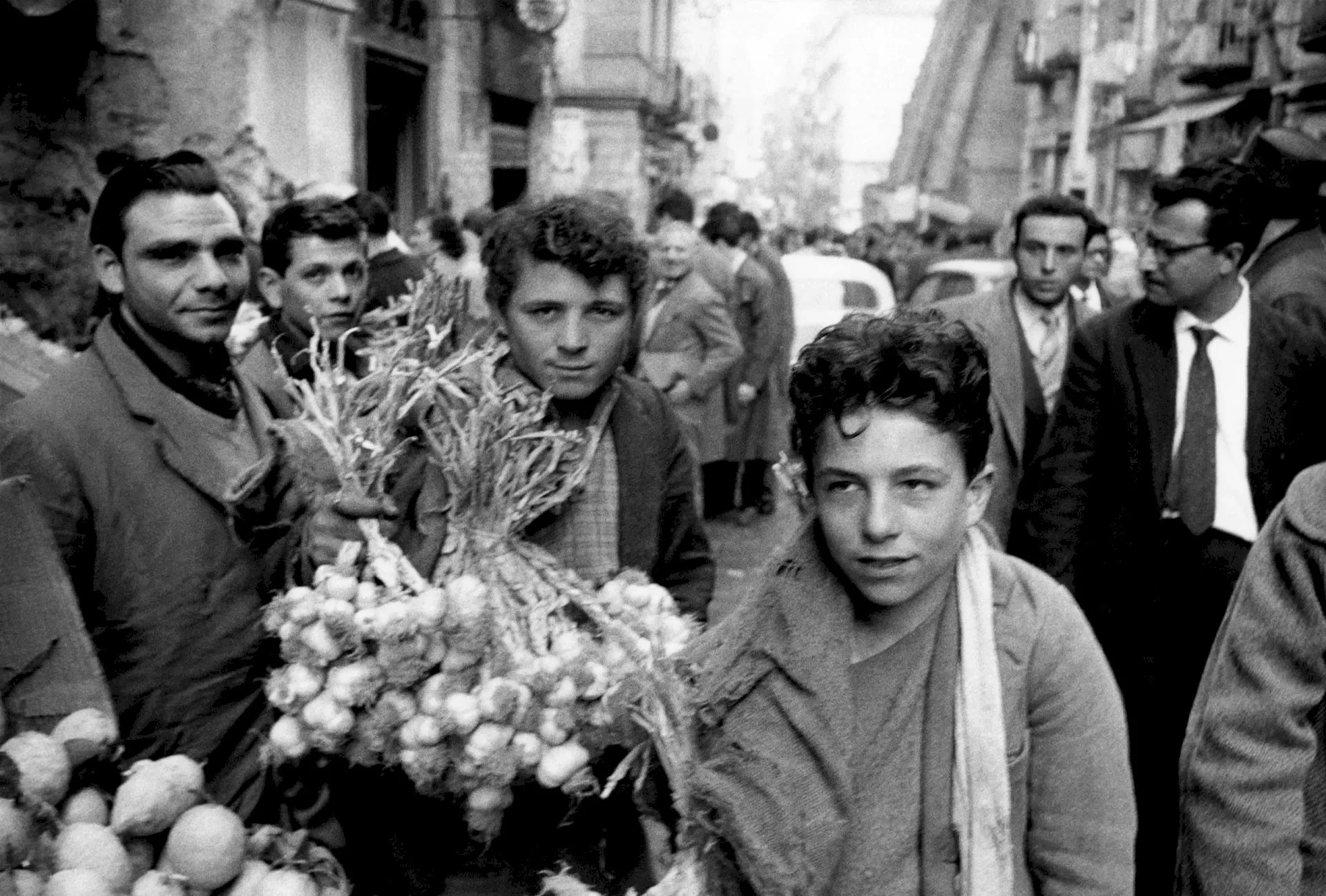 Black and white photo of boys and men in a street market with fruit, some boys in the foreground and men in suits in the background.