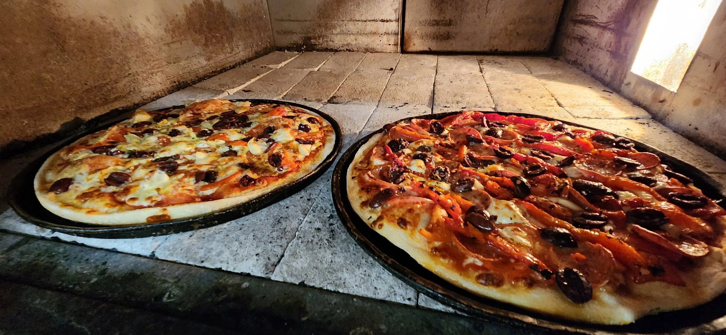 Two pizzas baking in a brick oven, one with black olives, cheese, and possibly chicken, and the other with pepperoni, cheese, black olives, and red bell peppers.