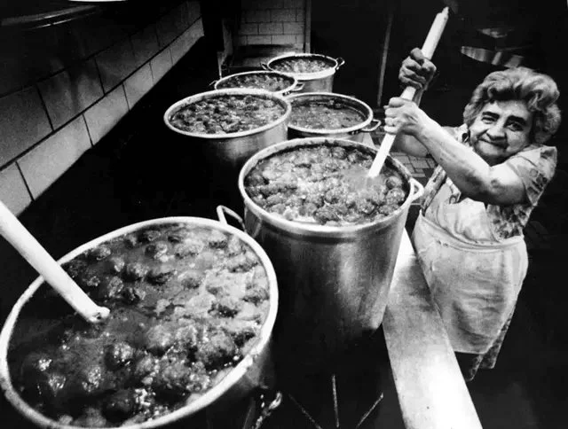 A woman stirring large pots of food in a kitchen.