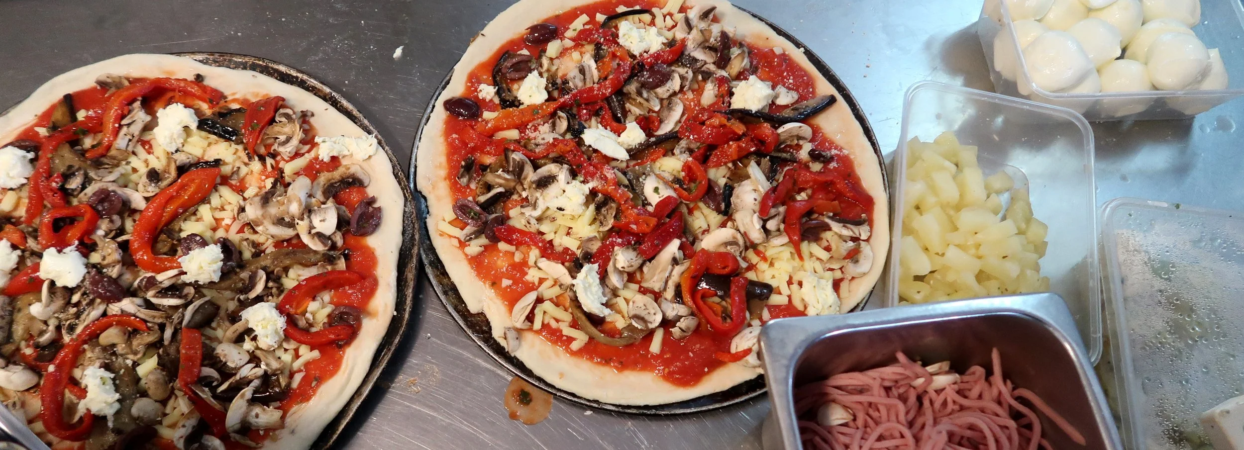 Two uncooked pizzas topped with cheese, mushrooms, red and green peppers, and black olives, sitting on a metal surface, with containers of cheese, ham, and other pizza toppings nearby.