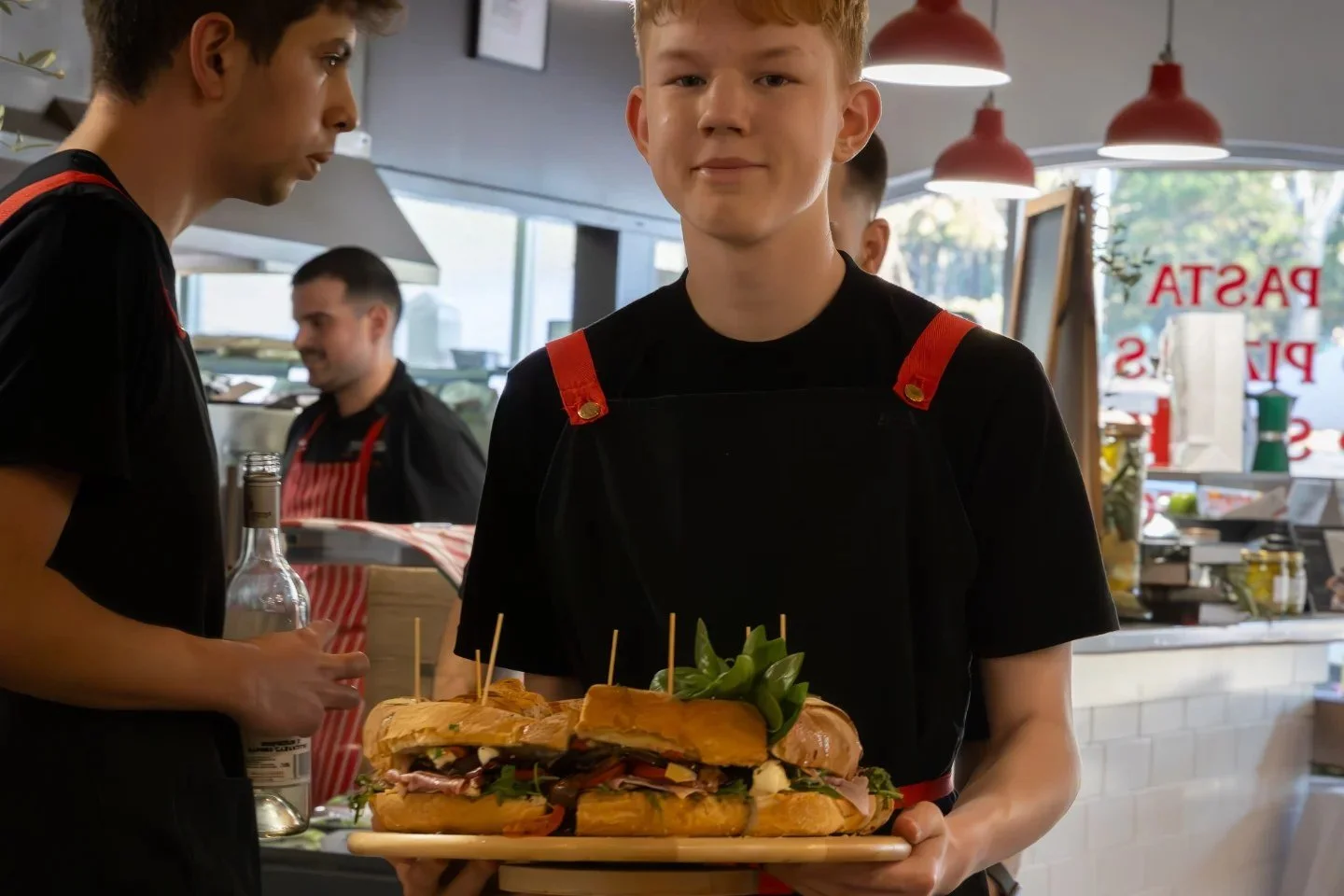 Young man holding a tray of sandwiches with various meats, cheese, and greens inside a cafe or restaurant.