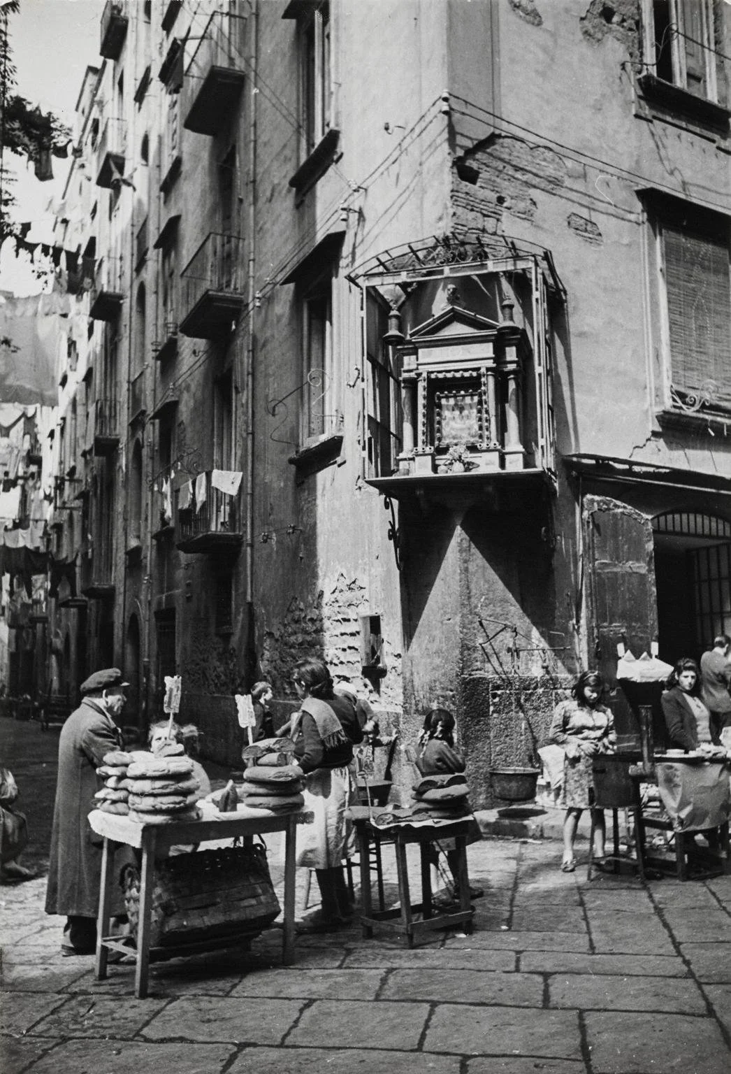 Street scene with people selling goods, some stacked on tables, in front of old buildings with laundry hanging from clotheslines.