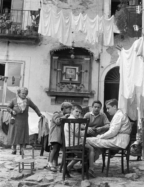 Group of children and woman sitting and standing in an outdoor courtyard with laundry hanging overhead and old buildings in the background.