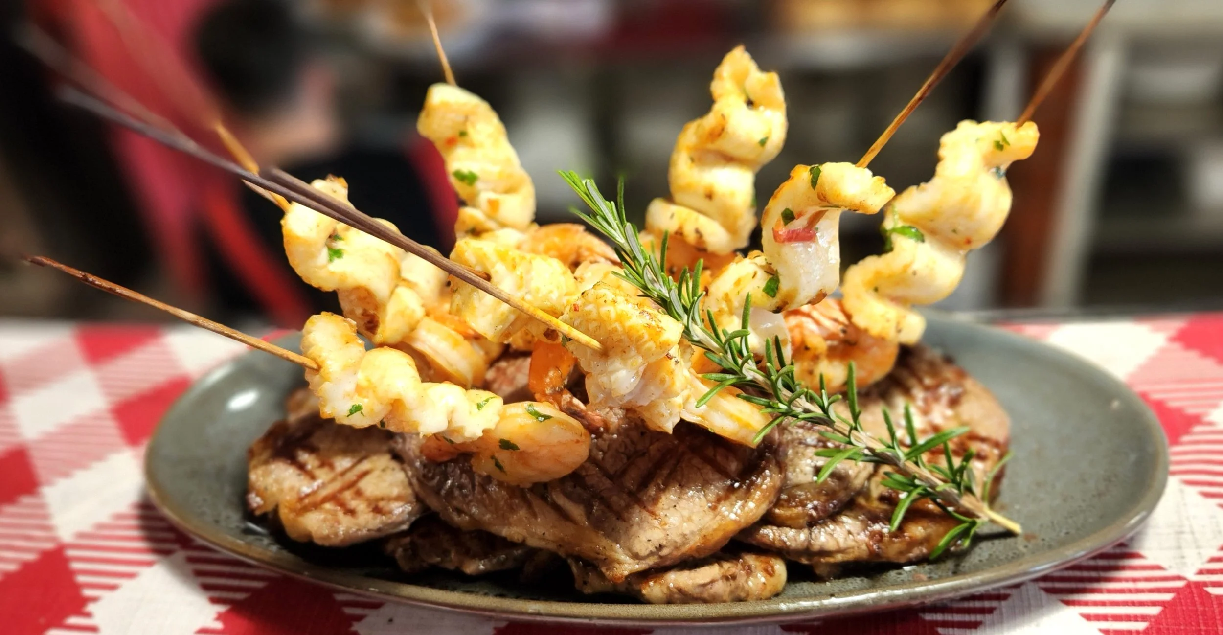 A dish featuring grilled steak topped with grilled shrimp skewers garnished with a sprig of rosemary, served on a gray plate on a red and white checkered tablecloth.