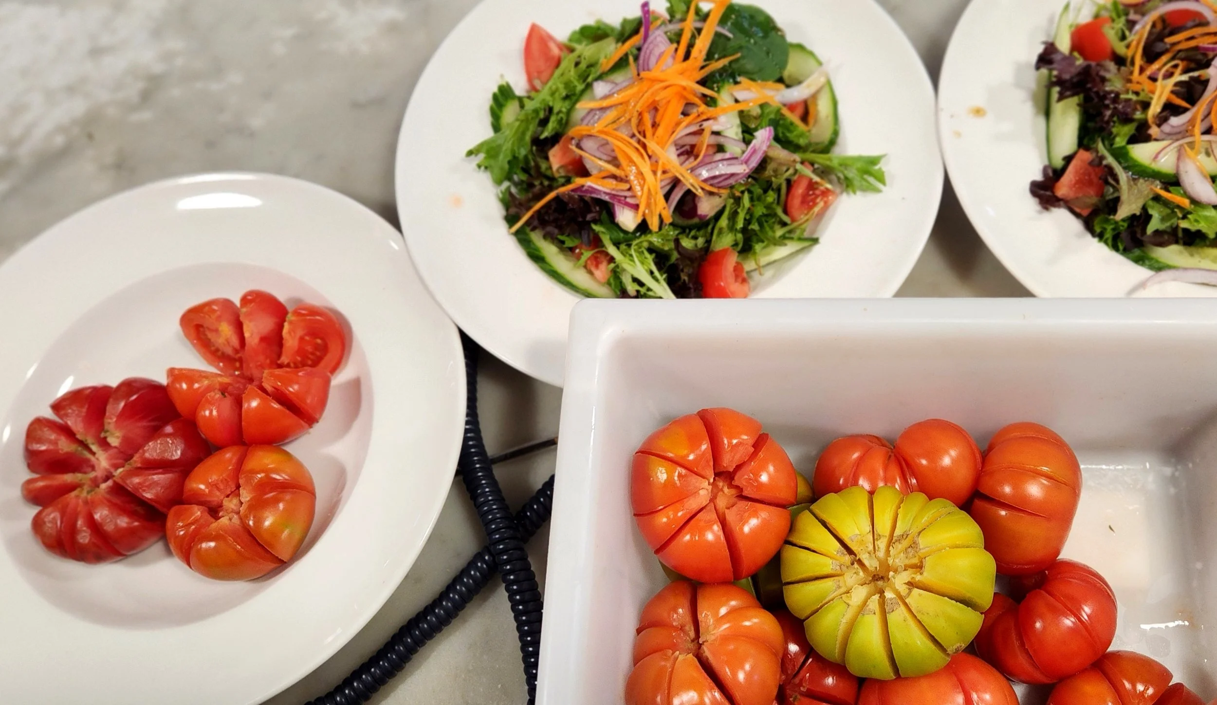 Assorted heirloom tomatoes and prepared salads with mixed greens, cherry tomatoes, shredded carrots, cucumbers, and onions on white plates.