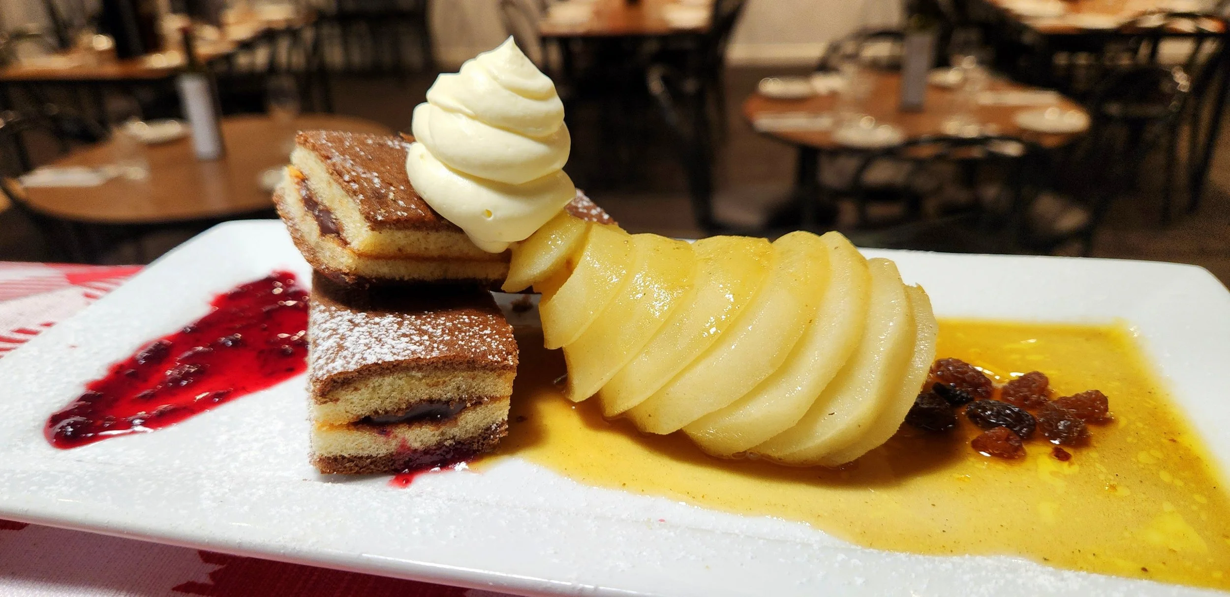 Plate with sliced pears, a dollop of whipped cream, slices of cake with berry filling, a red berry sauce, and a yellow sauce on a white rectangular plate.