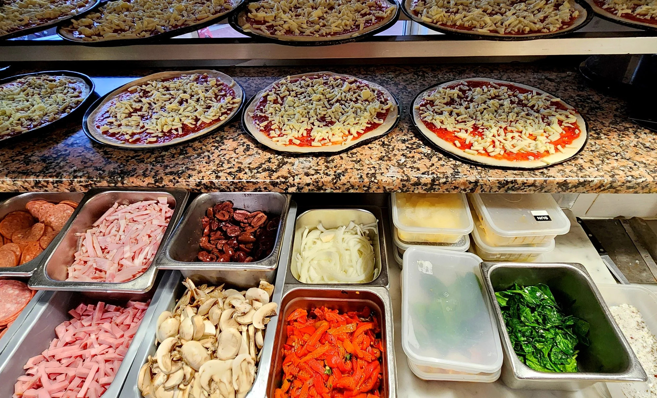 Pizzas with cheese and tomato sauce on a granite countertop, ingredients like mushrooms, onions, pepperoni, and various vegetables in metal containers.