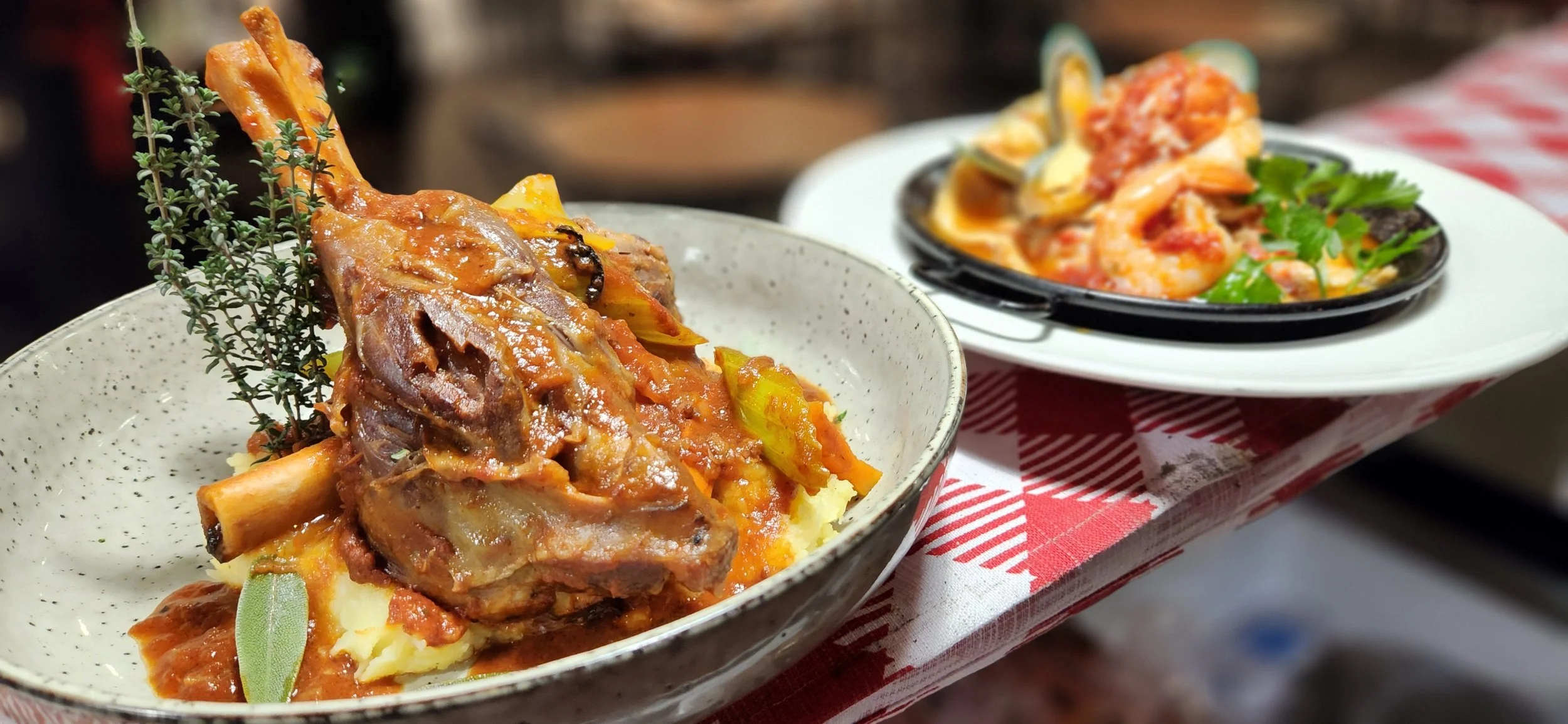 A ceramic bowl with a braised meat dish garnished with herbs, served over mashed potatoes. In the background, a plate of seafood pasta with shrimp and cilantro.