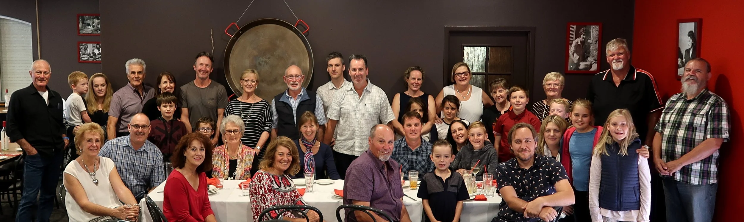 A large group of people, including adults and children, gathered in a restaurant for a celebration or family gathering, with tables set for a meal and decorations on the wall.