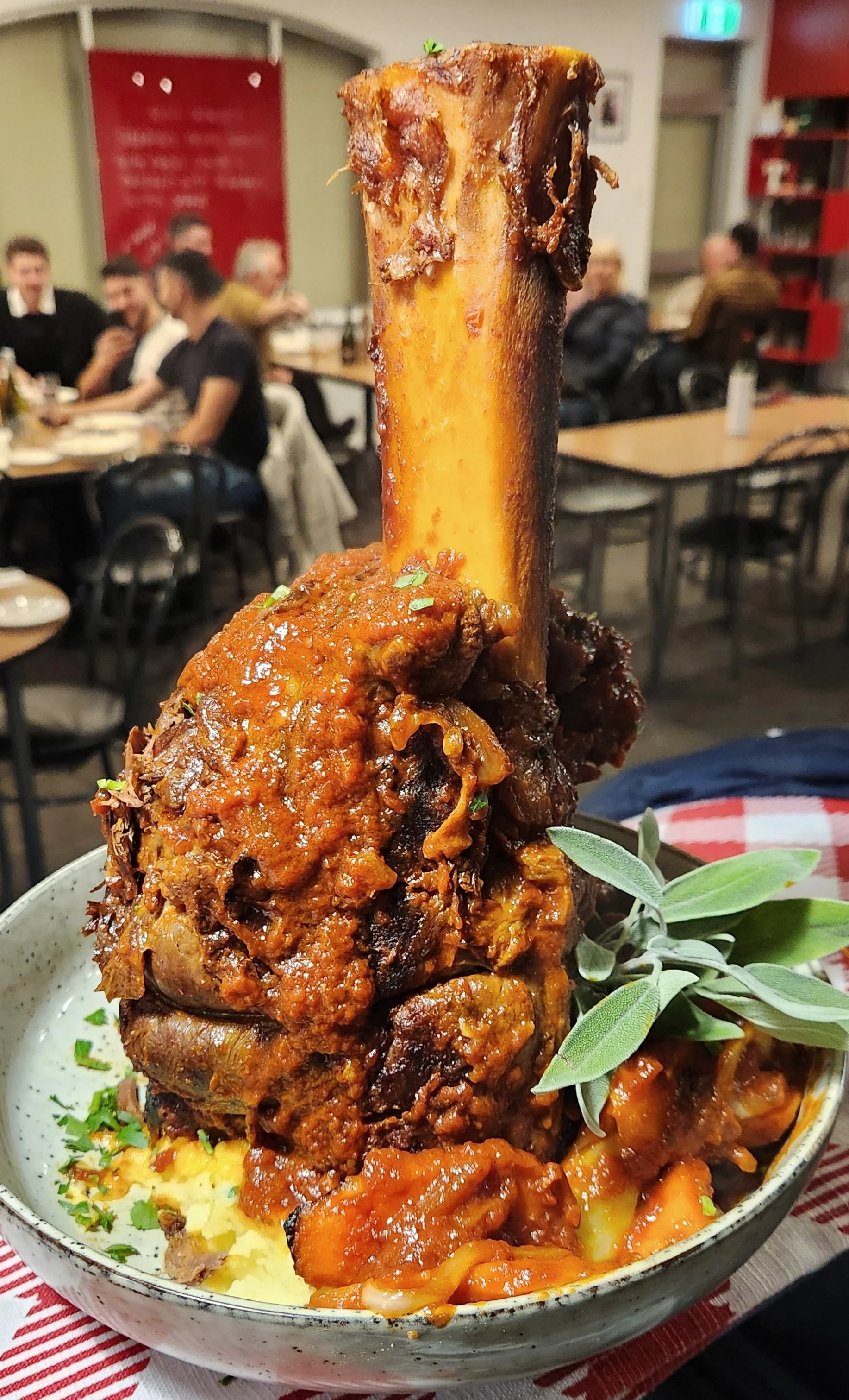 Close-up of a large beef shank on mashed potatoes, topped with tomato sauce and garnished with fresh herbs, served in a restaurant setting.