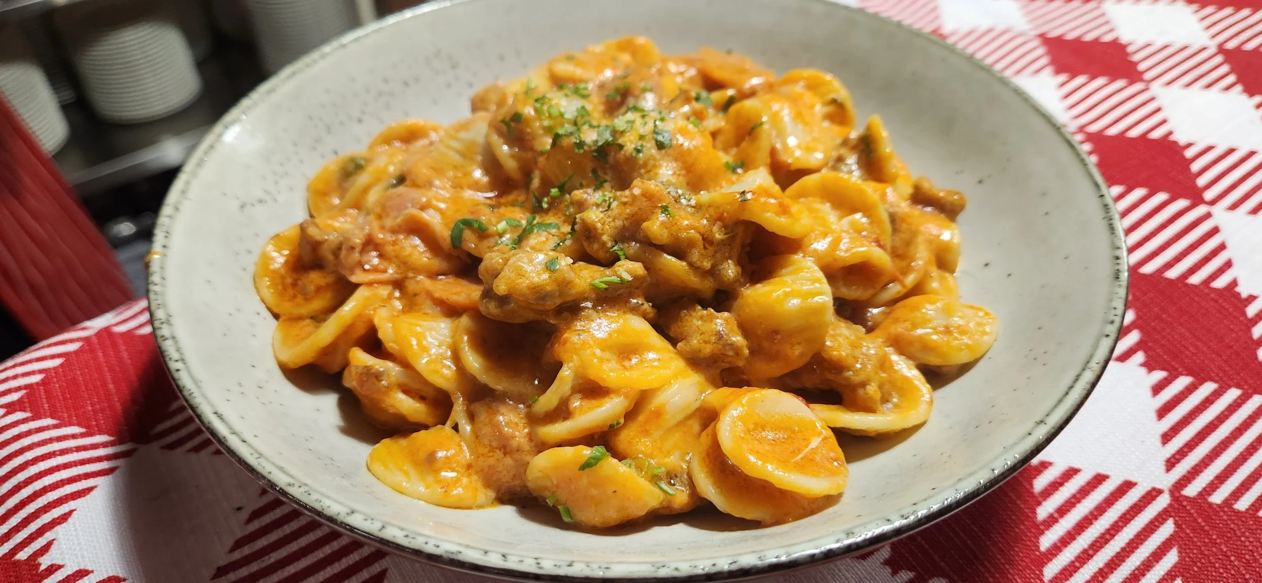 A bowl of cheesy pasta with ground meat, garnished with chopped herbs, on a red and white checkered tablecloth.