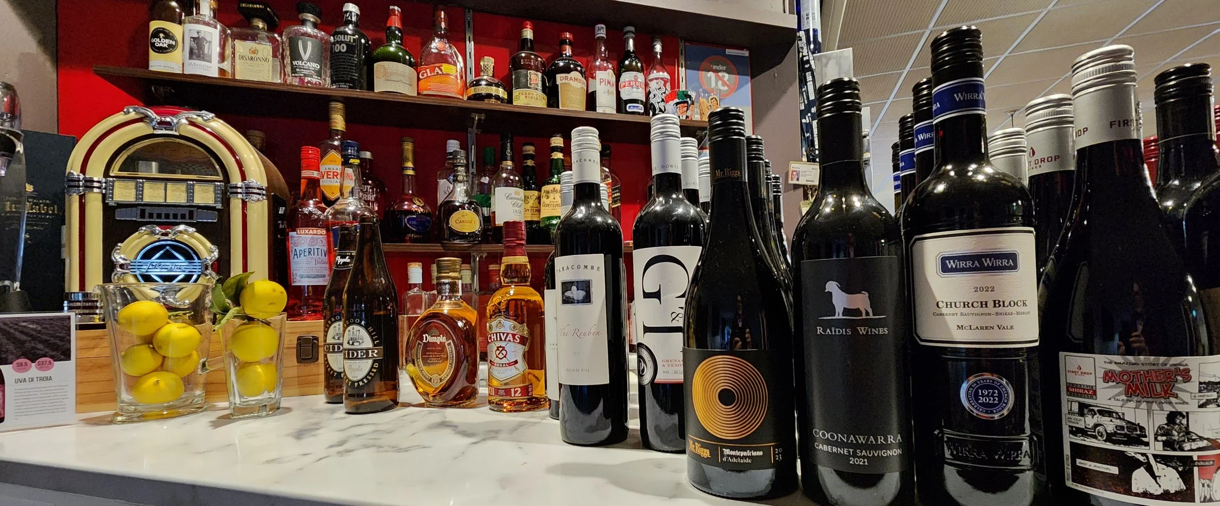 A selection of wine bottles and liquor bottles on a white marble bar counter with yellow lemons in a glass container, and a vintage jukebox in the background.