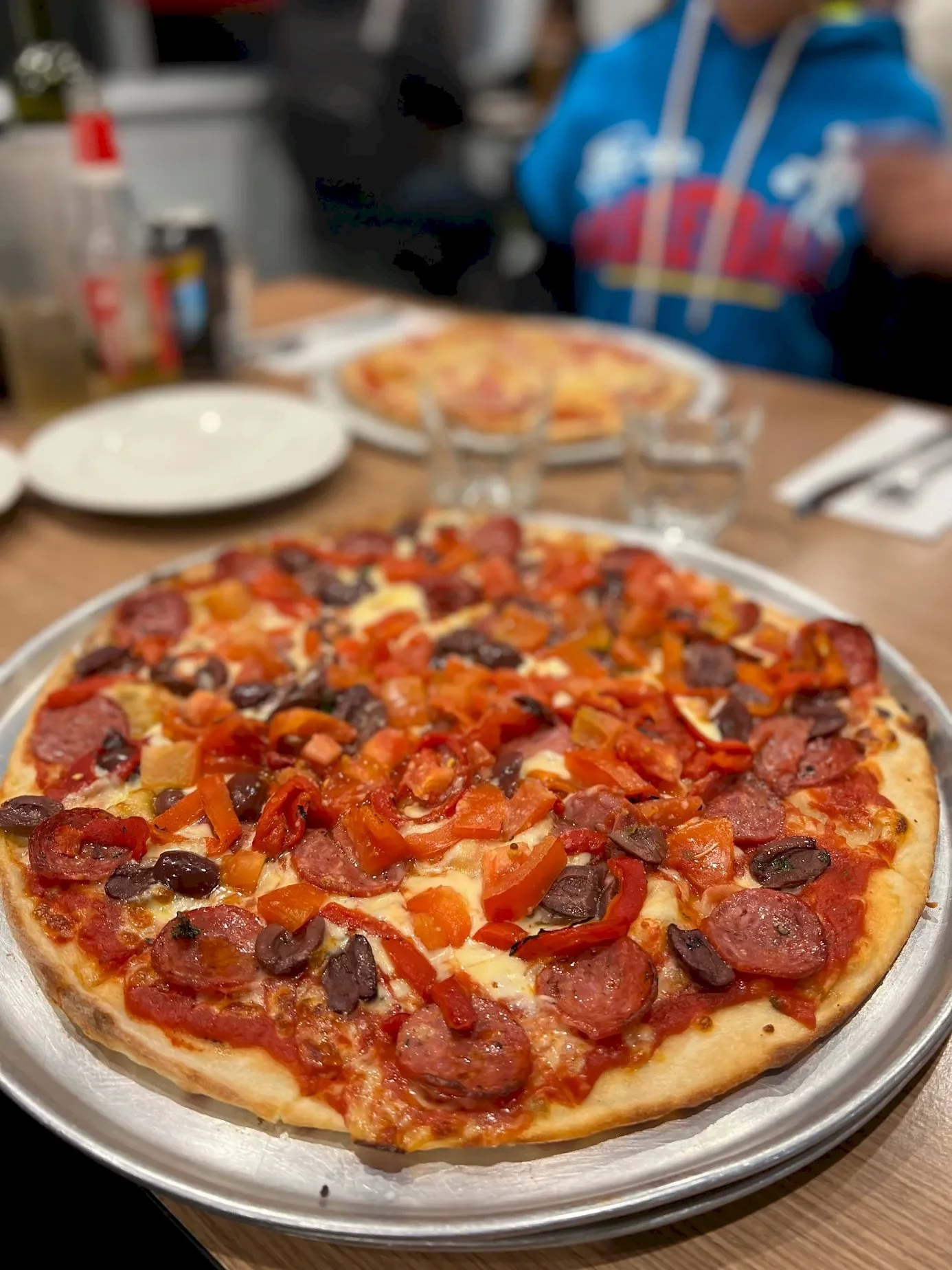 A pizza with pepperoni, black olives, and diced red and orange peppers on a silver tray at a dining table.