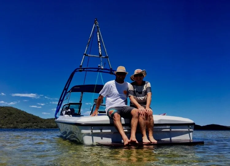 Two people sitting on the front of a white boat in clear water with a blue sky and distant hills in the background.