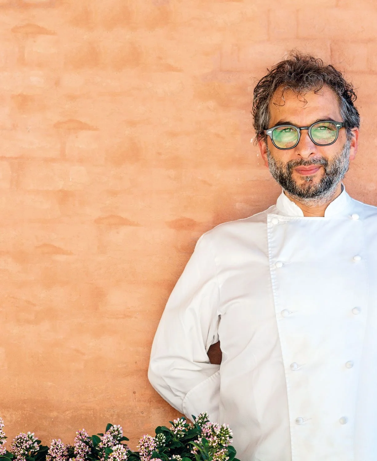 Uomo chef con occhiali, barba e capelli ricci, indossa una giacca bianca da cuoco, si trova davanti a una parete color terracotta e ai fiori rosa in primo piano.