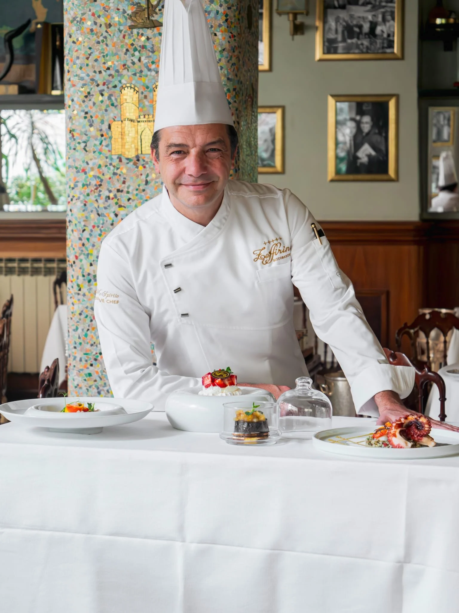 Chef sorridente indossa un cappello alto e una giacca bianca, seduto davanti a un tavolo apparecchiato con vari piatti gourmet, decorato con piatti di cibo elaborato, in un ristorante con pareti adornate di quadri.