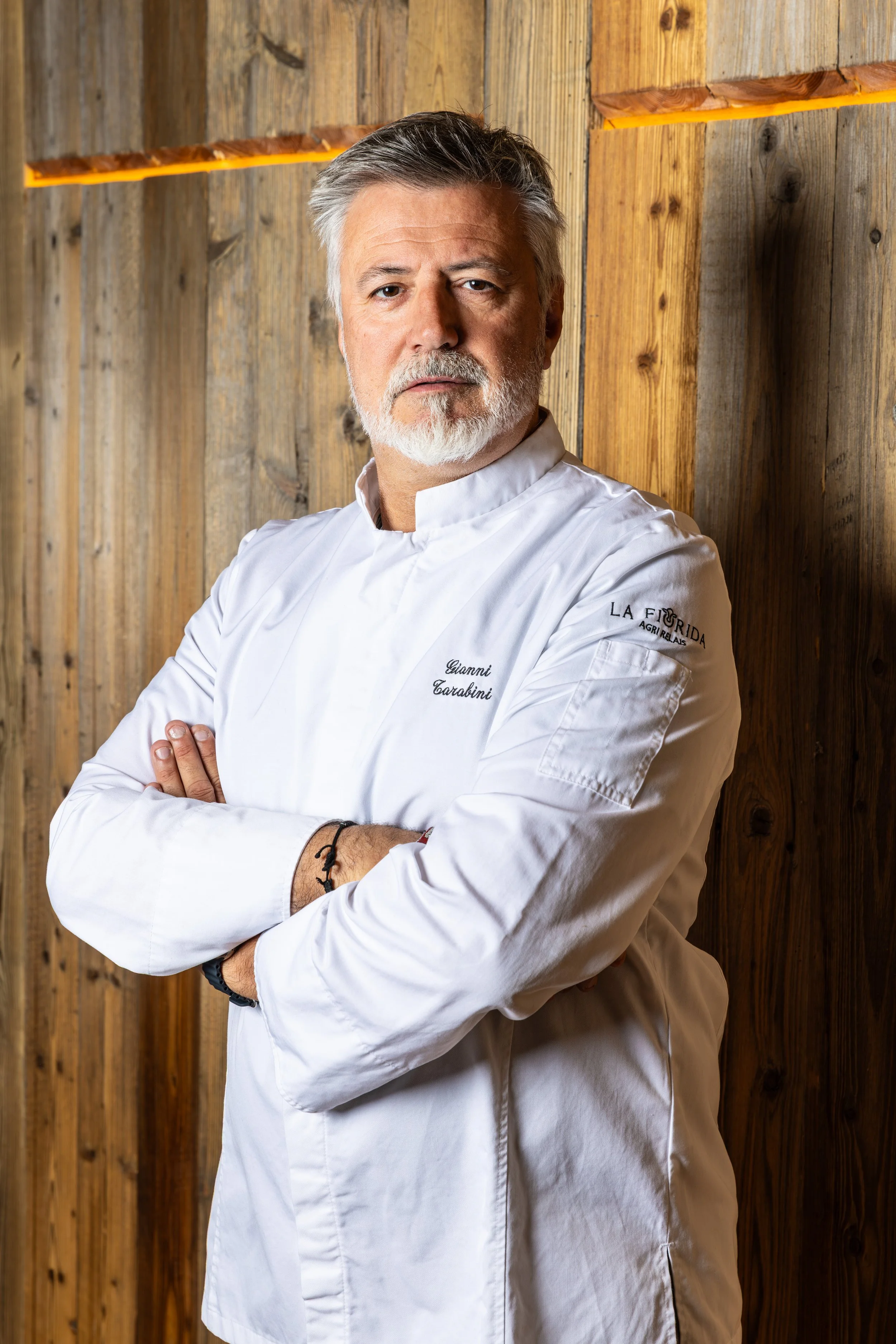 Uno chef con barba e capelli grigi, indossa una giacca da chef bianca con il nome 'Gianni Carabini' ricamato e posizionato con le braccia incrociate davanti a uno sfondo di legno.