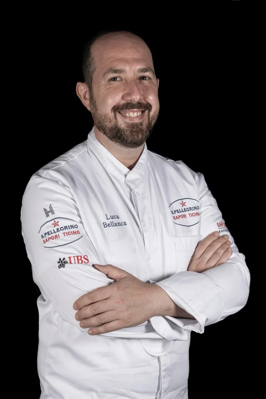 Uno chef sorridente indossa una giacca bianca con vari loghi, inclusi S. Pellegrino, UBS e SAPORI TICINO, e si trova contro sfondo nero.