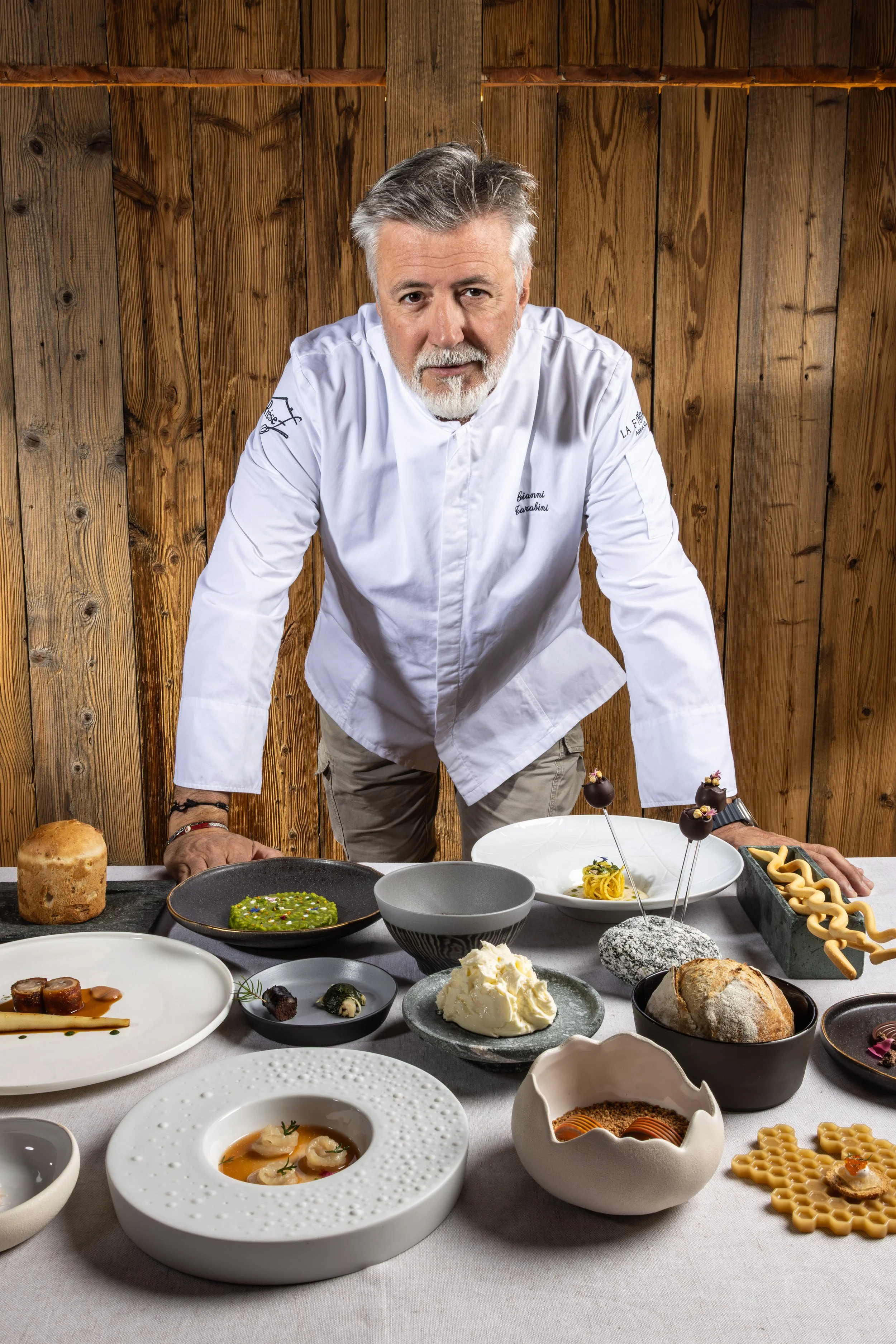 Uno chef mascherato con grembiule bianco si inchina davanti a una tavola con piatti di vari cibi, tra cui zuppe, pane, dolci e antipasti, in un ambiente rustico con pareti di legno.
