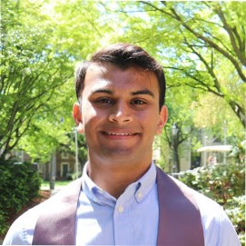 A young man smiling outdoors with green trees and a building in the background.