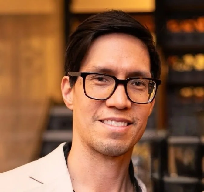 A man with dark brown hair, glasses, and a slight smile in an indoor setting with blurred warm-colored background.