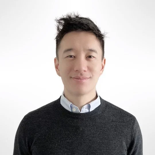 A young man with short black hair and a slight smile, wearing a white collared shirt and a dark gray sweater, standing against a plain white background.