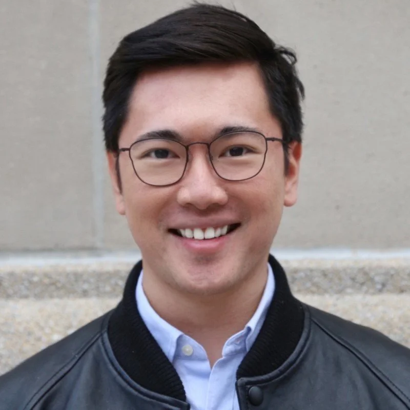 Young man with glasses smiling outdoors, wearing a leather jacket and a button-down shirt.