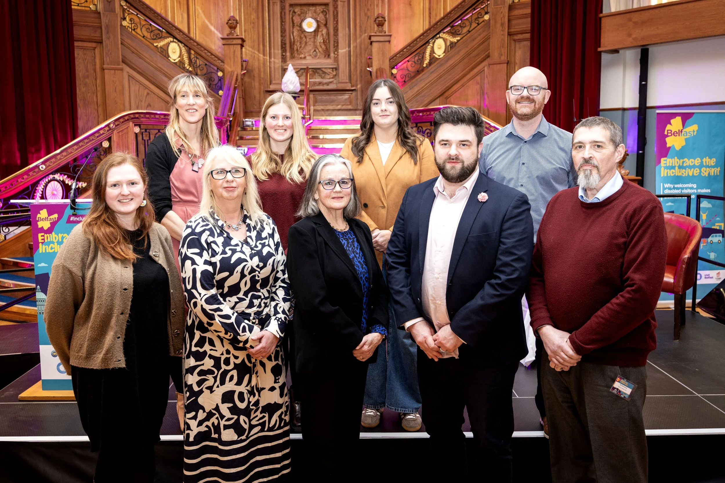 Group of ten people posing indoors on a stage with wooden paneling, stairs, and bright lights, at a conference or event in Belfast.