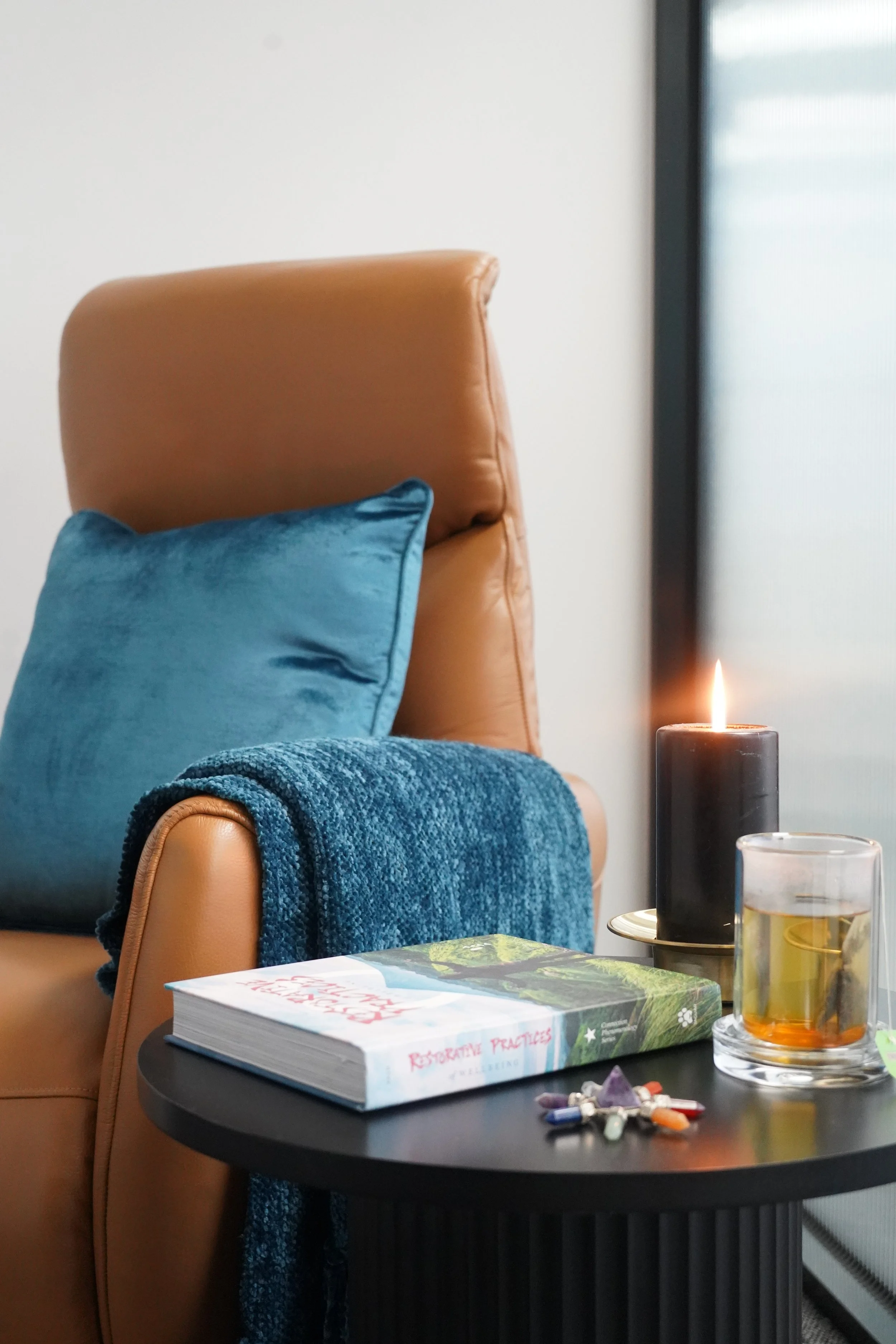 A black side table with a large book titled 'Restorative Practices,' two candles (one black and one pillar), a glass of whiskey, and assorted pills or capsules. A tan leather armchair with a teal pillow and blanket, next to a window with natural light.
