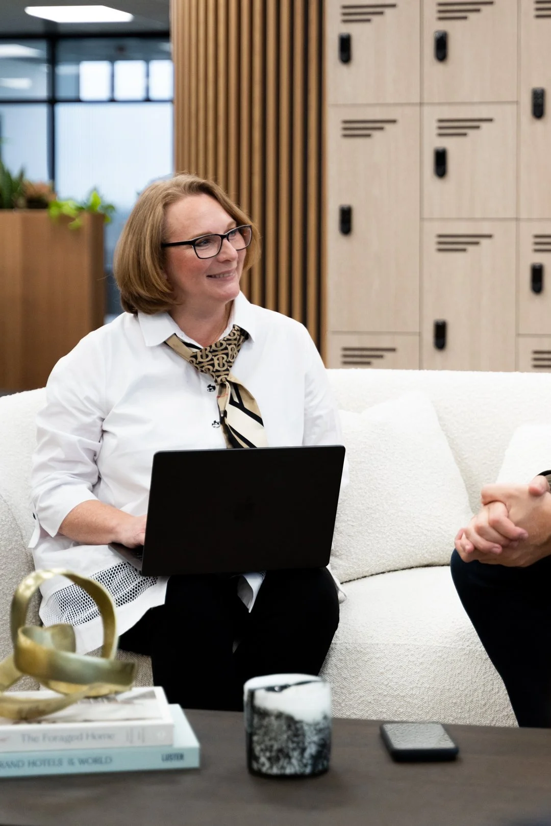 A woman with glasses and a patterned scarf around her neck sitting on a cream-colored sofa with a laptop on her lap, in a modern office lobby setting, smiling and talking to a person whose hands are visible.