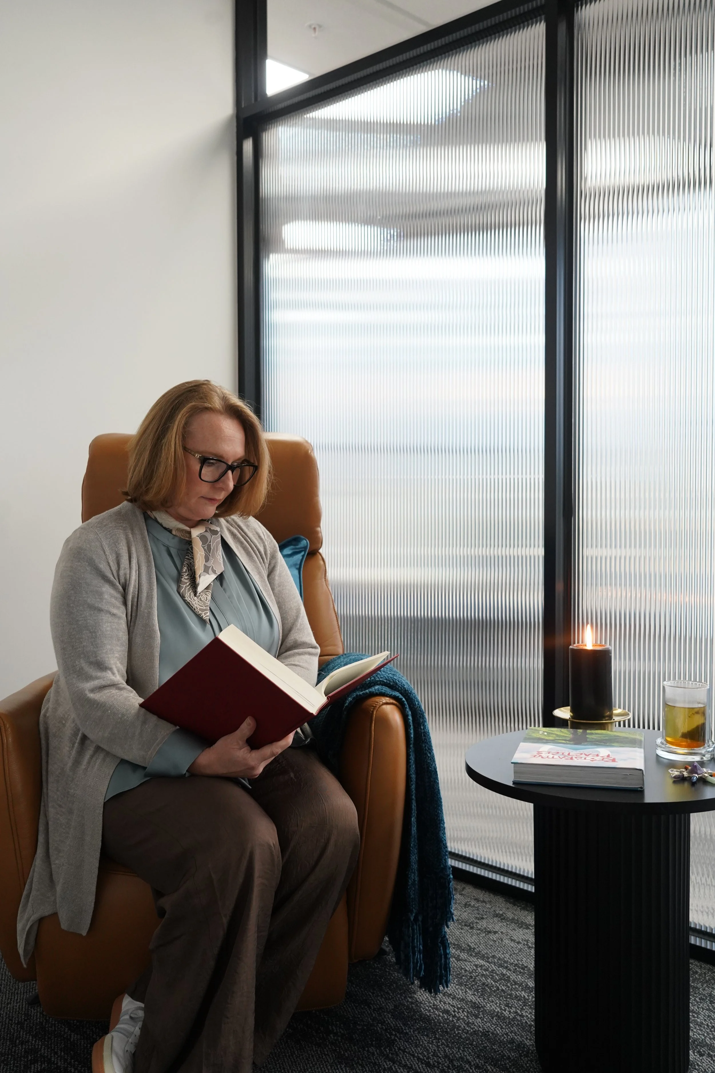 A woman with glasses, wearing a light gray cardigan and brown pants, is sitting in a tan leather chair reading a red book. Next to her, on a black side table, there is a lit black candle, a book, a glass of beer, and some keys.