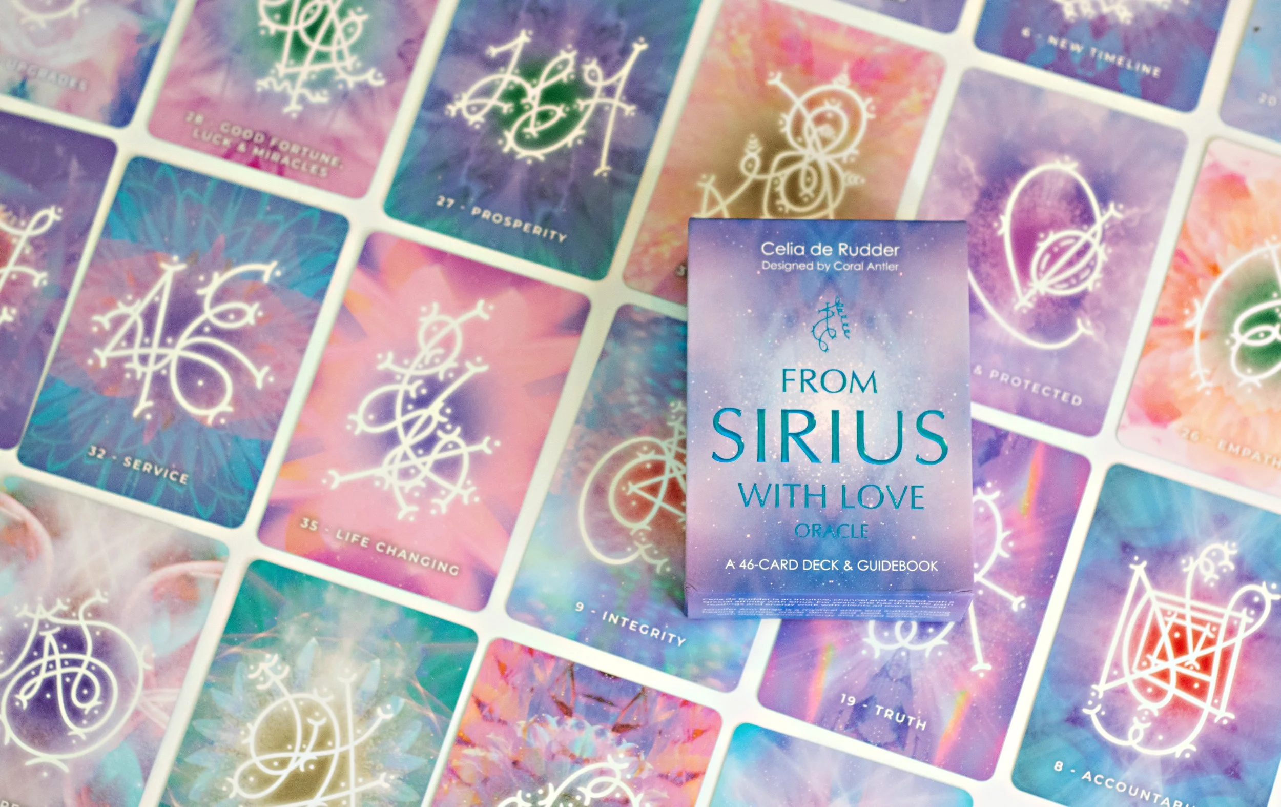 A deck of tarot or oracle cards titled 'From Sirius with Love', designed by Celia de Rudder, placed on a colorful spread of similar cards featuring astrological or mystical symbols and words like 'Service', 'Life Changing', 'Integrity', and 'Truth'.