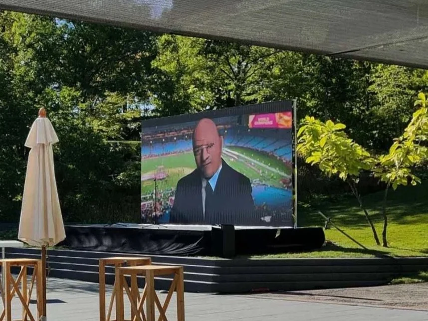 Outdoor outdoor screening setup showing a large screen with a severe-looking man in a suit on it, surrounded by trees and outdoor furniture.