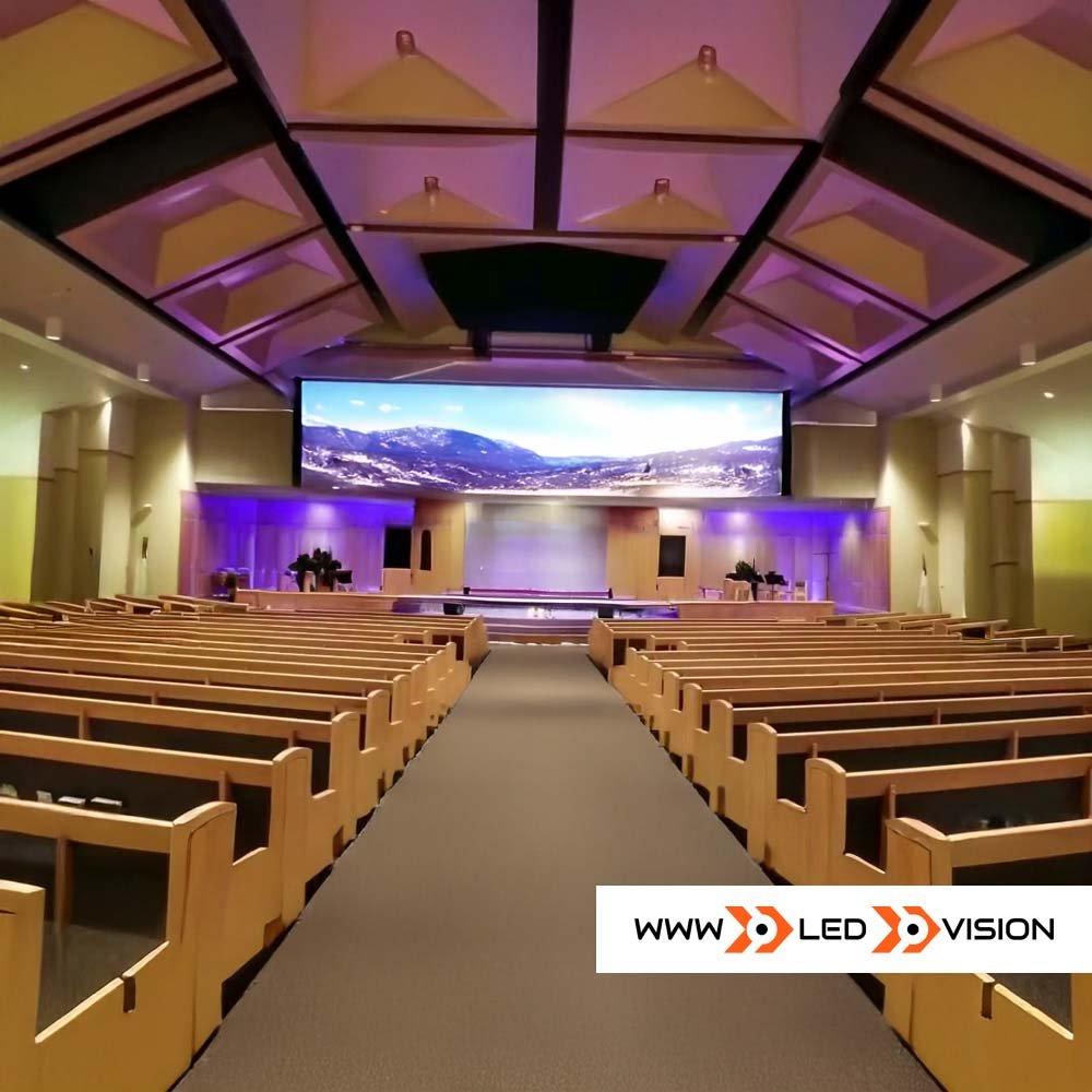 An auditorium with rows of wooden pews facing a stage with a large curved screen displaying a mountain landscape. The ceiling has geometric shapes and purple lighting accents.