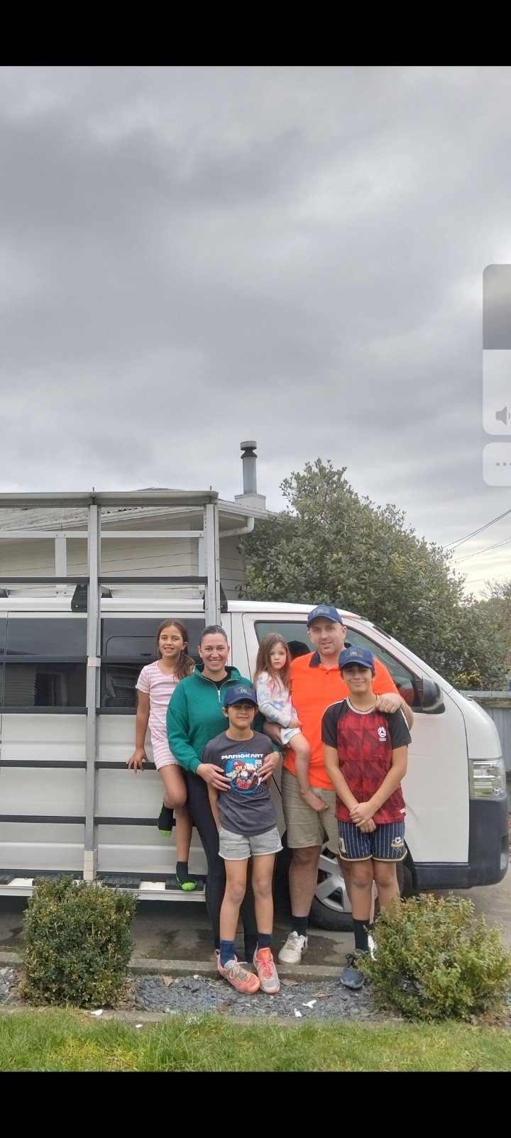 Family of six posing in front of a white van with a ladder on its side, outdoors on a cloudy day. There are trimmed bushes and a house with a chimney in the background.