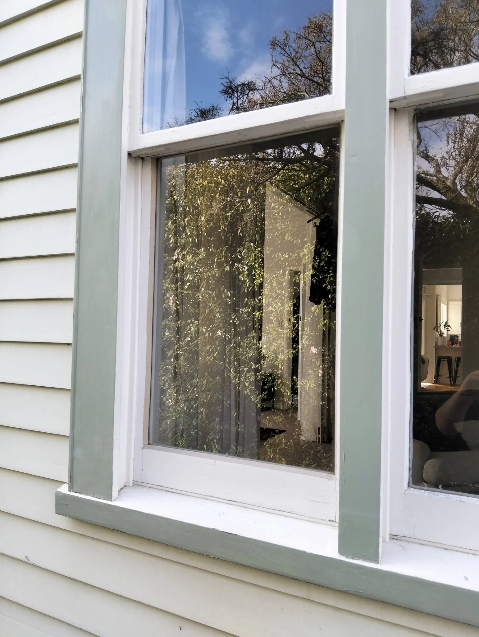 Close-up of a house window with a white frame and light green trim, reflecting blue sky and trees outside; interior visible through glass.