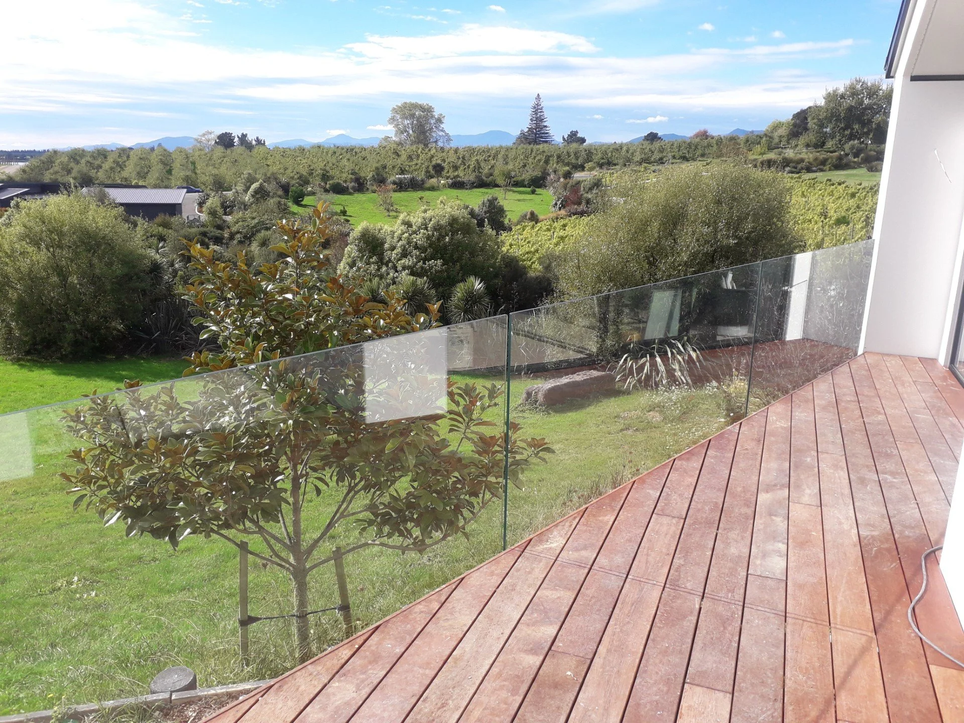 View from a wooden deck overlooking a lush green yard with trees, shrubs, and distant rolling hills under a partly cloudy sky. Frameless glass balustrade for fall protection and at the same time not obstructing the view.
