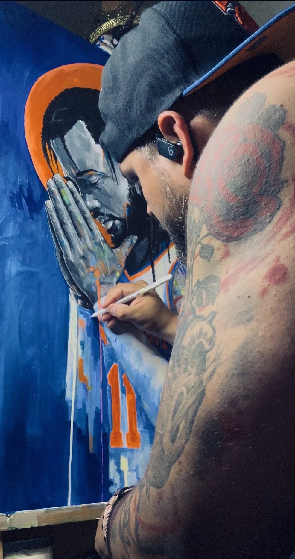 An artist with tattoos on his arm, a baseball cap, and wireless earbuds is painting a portrait of Jalen Brunson with a  prayer, eyes closed, and a vivid orange halo around his head on a blue background.