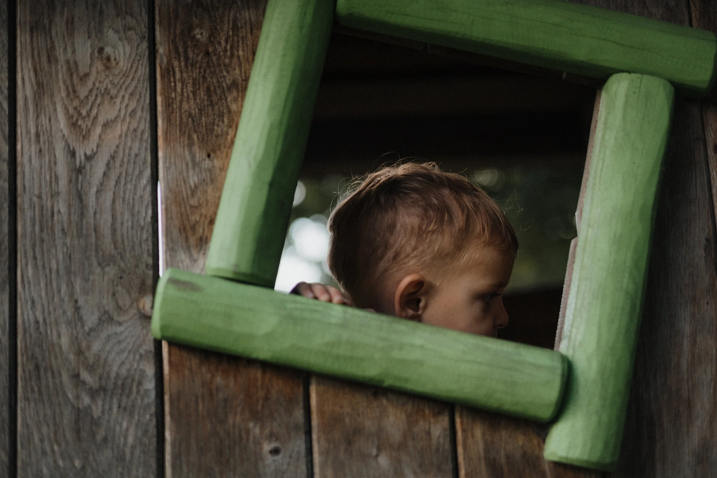 Nahaufnahme eines Jungen, der durch ein quadratisches Fenster in einer Holzhütte schaut, das mit grünen Holzstücken umrissen ist.