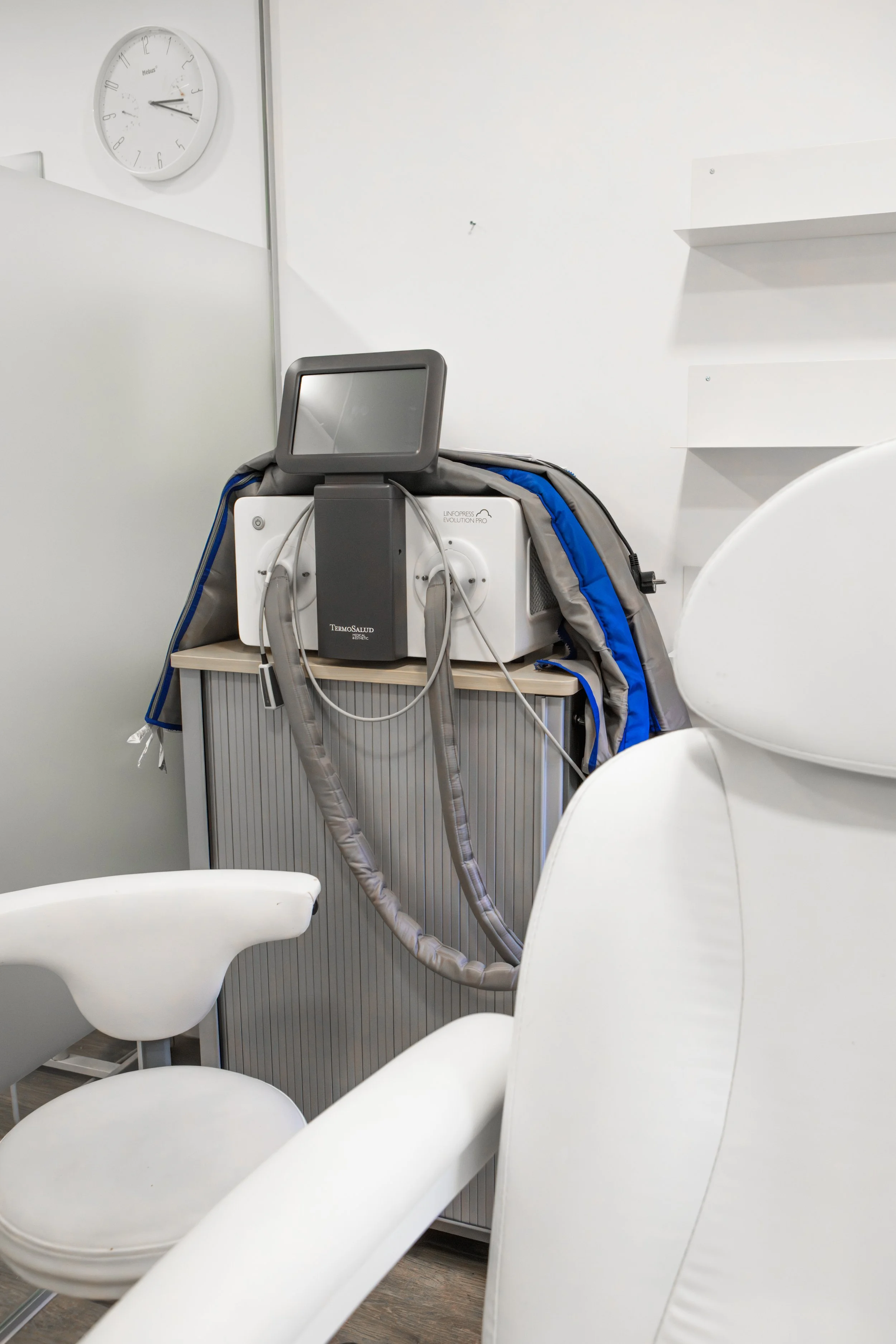 Medizinisches Gerät in einem weißen Behandlungsraum mit weißen Stühlen und einer Uhr an der Wand.