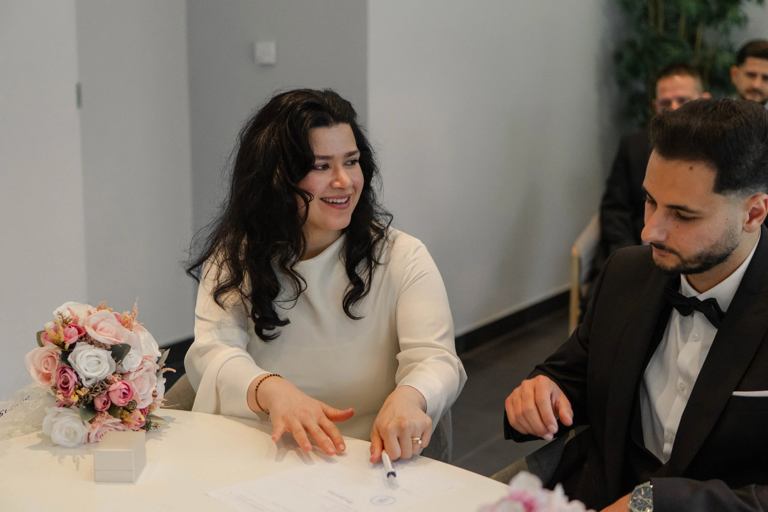 Eine Frau mit dunklen, lockigen Haaren und einem weißen Kleid spricht mit einem Mann in einem schwarzen Anzug und Fliege bei einer Heiratszeremonie. Auf dem Tisch liegt ein Blumenstrauß aus rosa und weißen Rosen, und es sind einige Dokumente sichtbar