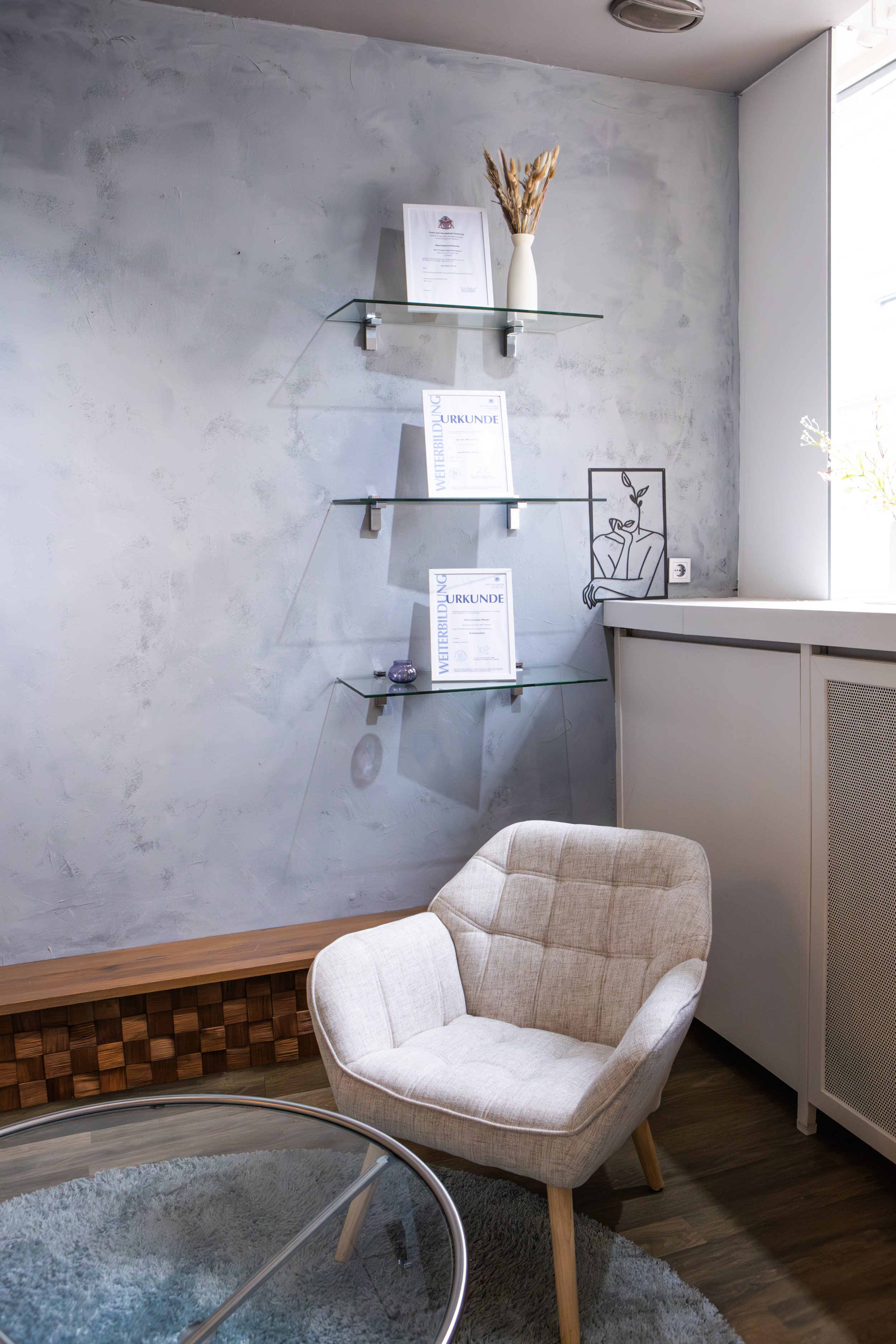 Ein modernes Wohnzimmer mit einer weißen, gepolsterten Sessel, einem runden Glastisch, einem grauen Wand mit eingehängten Glasregalen, die mit Urkunden und einer Vase mit trockenen Pflanzen dekoriert sind, einem Fenster, das Tageslicht hereinlässt, u