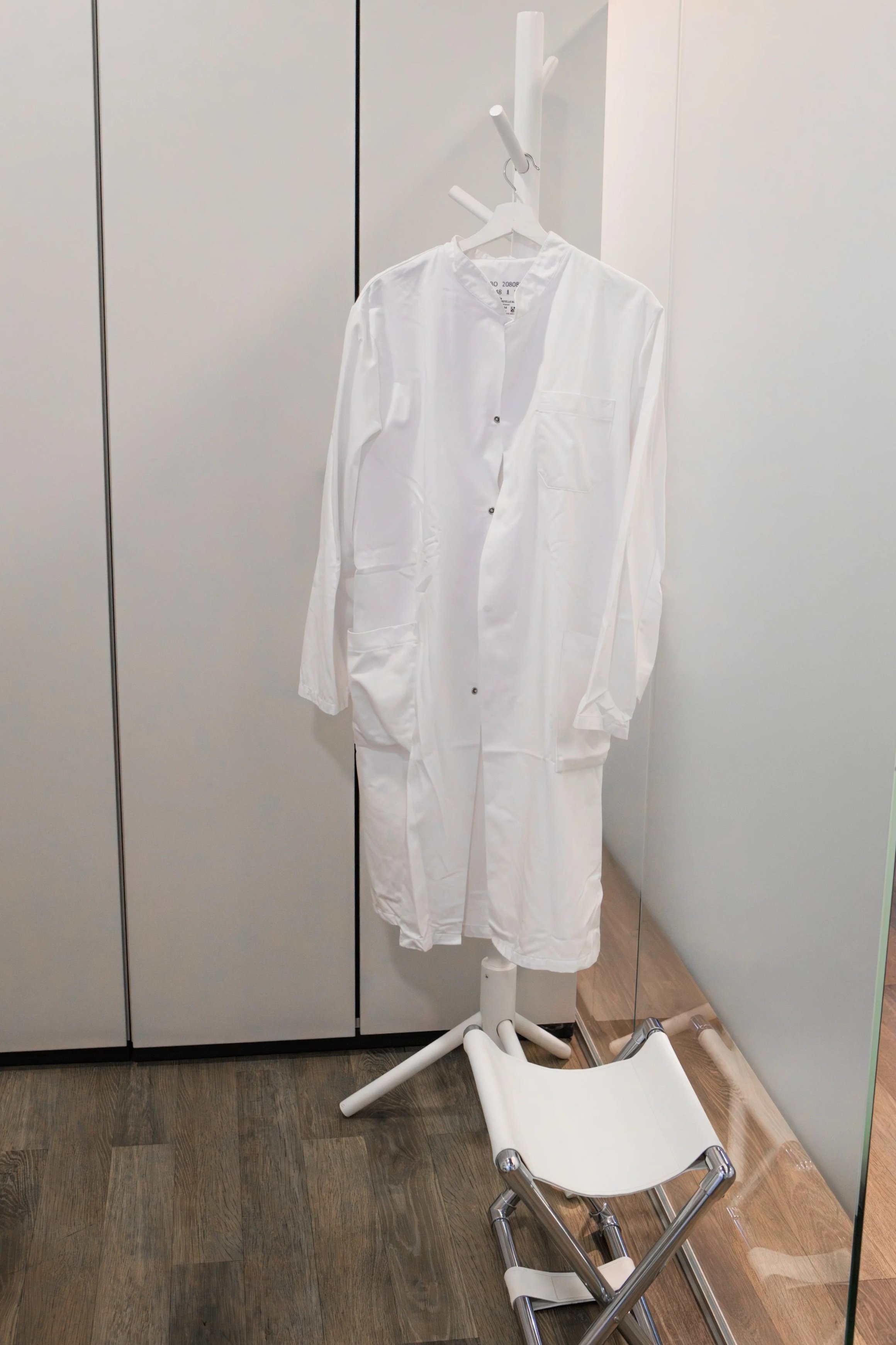 Ein weißer Kittel hängt an einem Kleiderständer in einem Raum mit weißen Wänden und Holzfußboden.