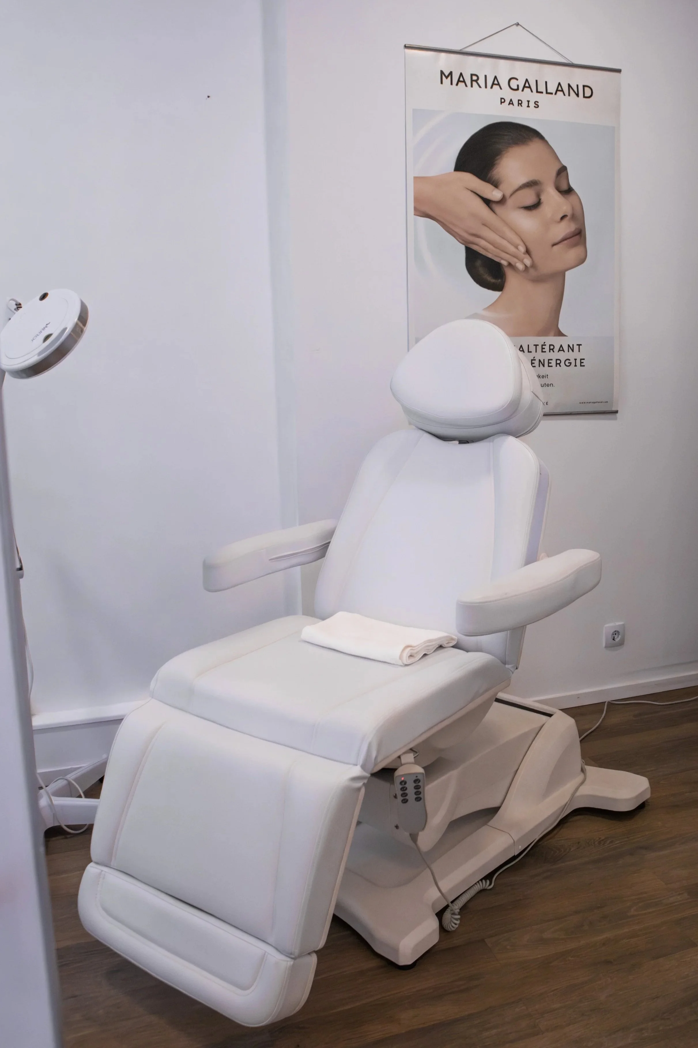 Ein weißer Behandlungsstuhl in einem Kosmetikstudio mit einem Poster an der Wand, das eine Frau mit geschlossenen Augen zeigt, die eine Gesichtsmassage erhält. Der Raum hat Holzfußboden und weiße Wände.