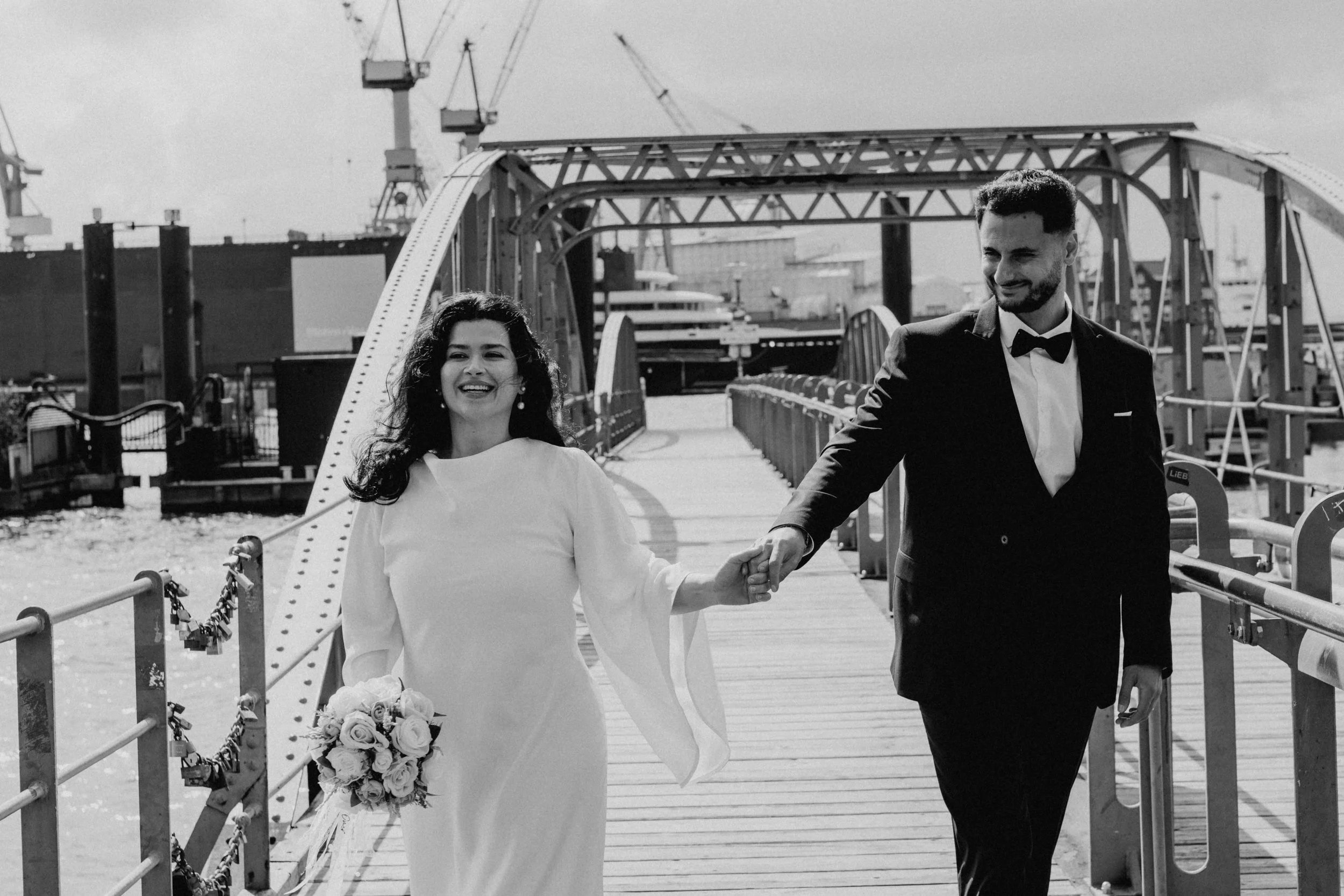 Ein glückliches Hochzeitspaar, ein Mann in Smoking und eine Frau in einem weißen Hochzeitskleid, hält sich an einem Steg am Wasser fest, im Hintergrund sind Schiffe und Kräne zu sehen, schwarz-weiße Aufnahme.