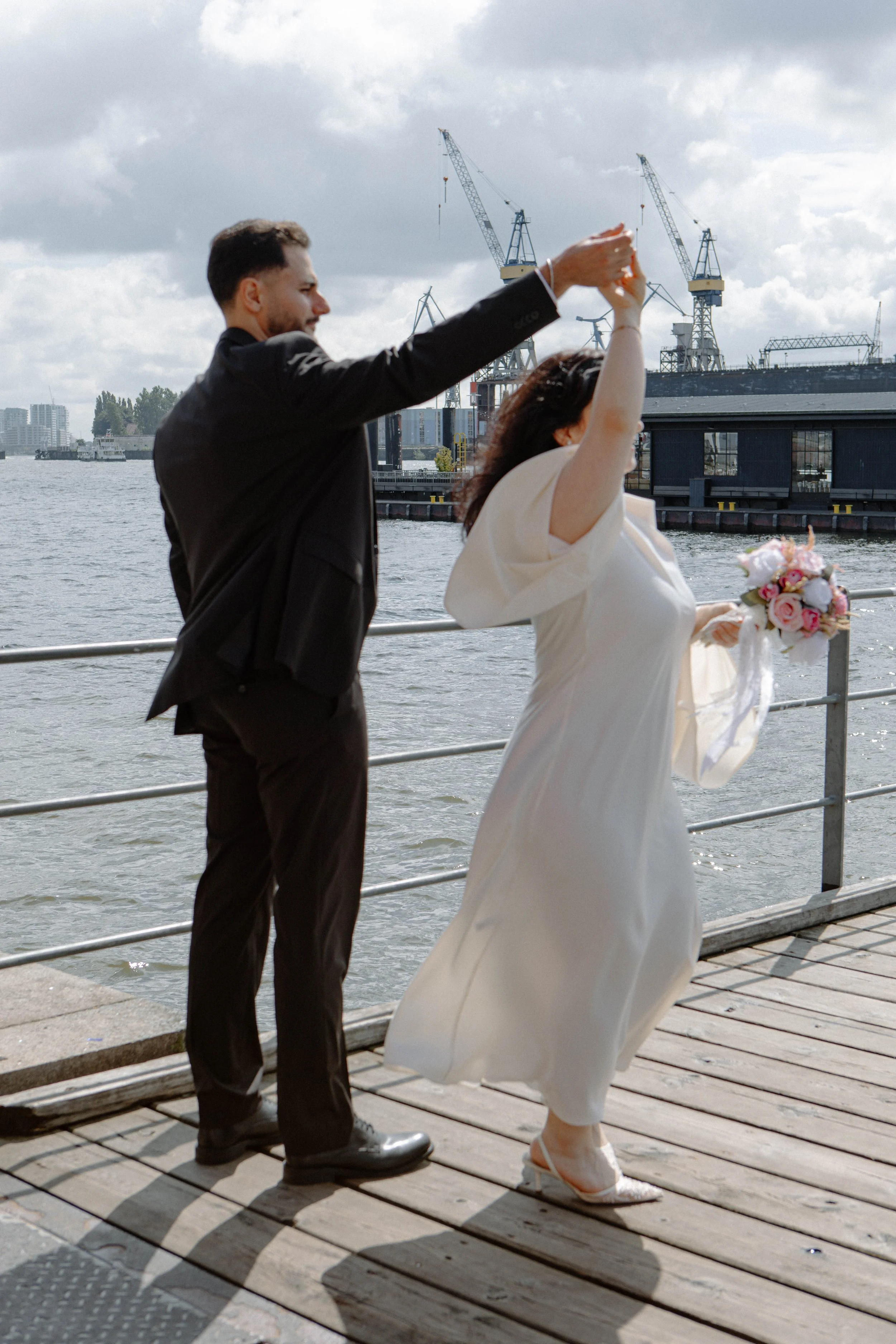 Ein Paar bei einer Hochzeit am Wasser, tanzen auf einem Steg, mit Stadt und Baukränen im Hintergrund.