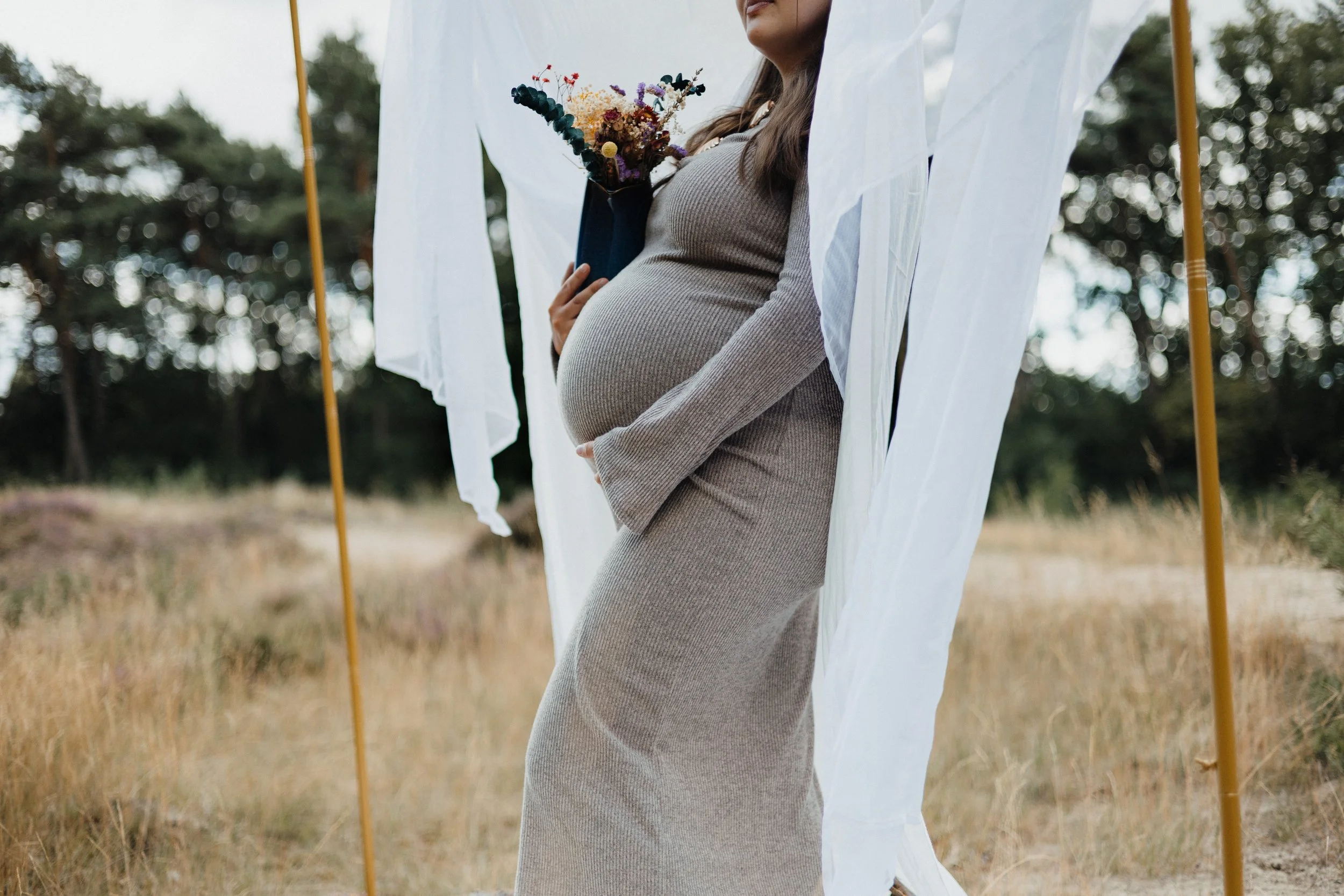 Schwangere Frau in einem langen, beigefarbenen Kleid, die ein Bouquet hält, steht in einer Outdoor-Umgebung mit weißen Stofftüchern und einem Rahmen aus Holzstangen.